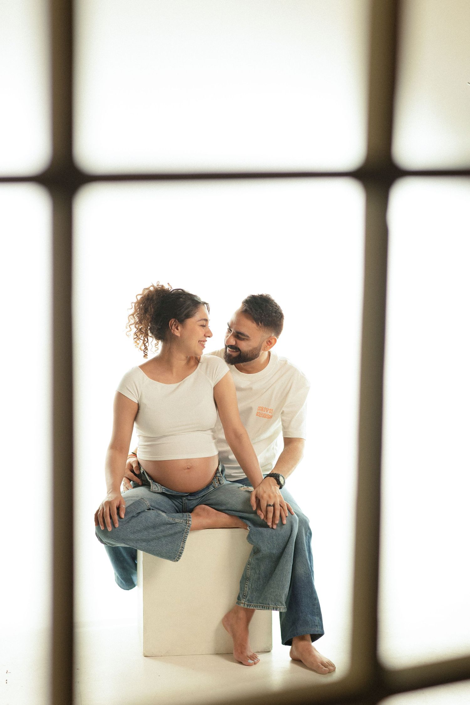 Couple in white shirts and jeans sitting together in intimate maternity photoshoot with window lighting.
