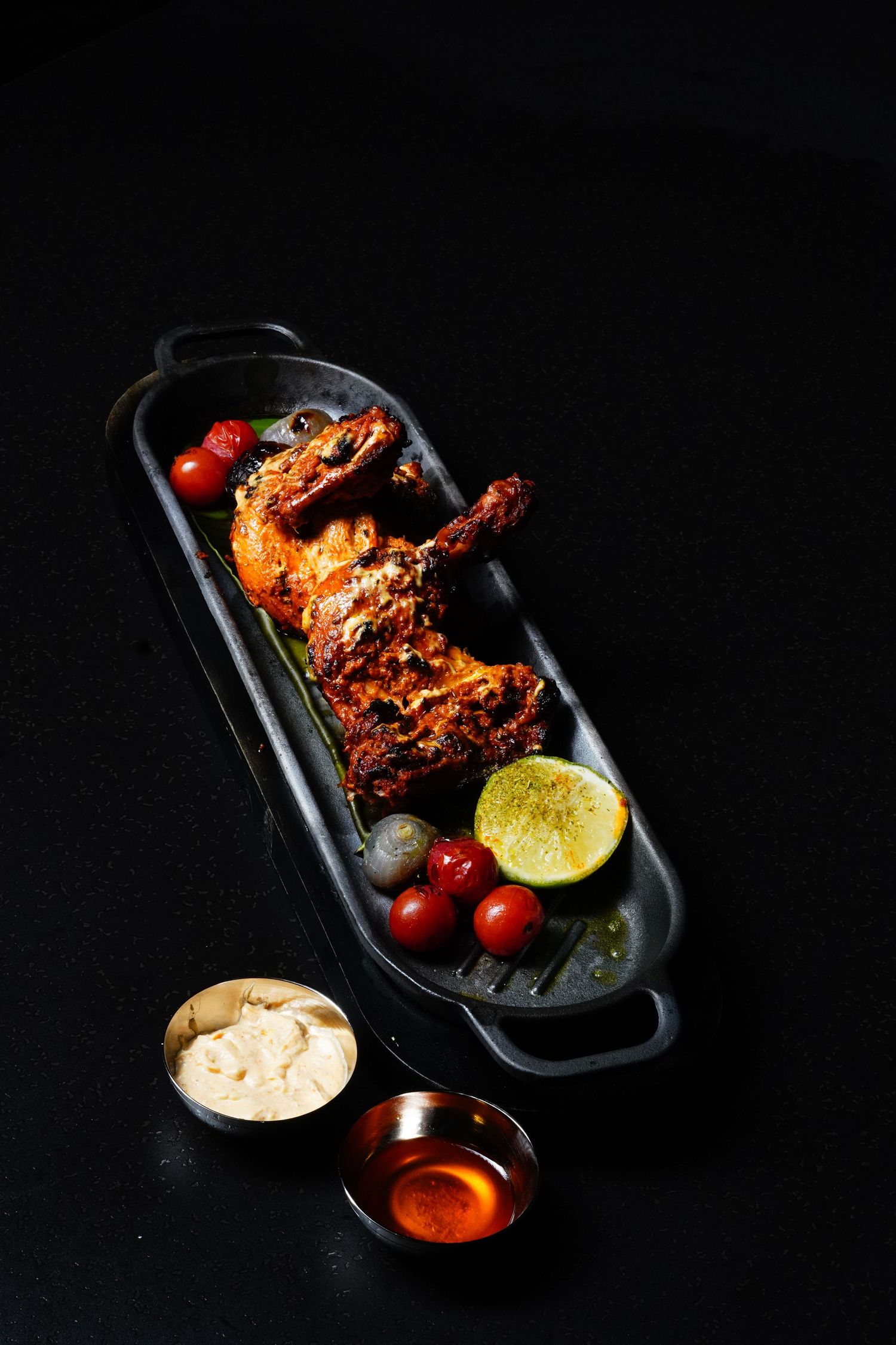 Grilled spicy chicken wings served with lime wedges, cherry tomatoes, and dipping sauces on black plates against dark background.