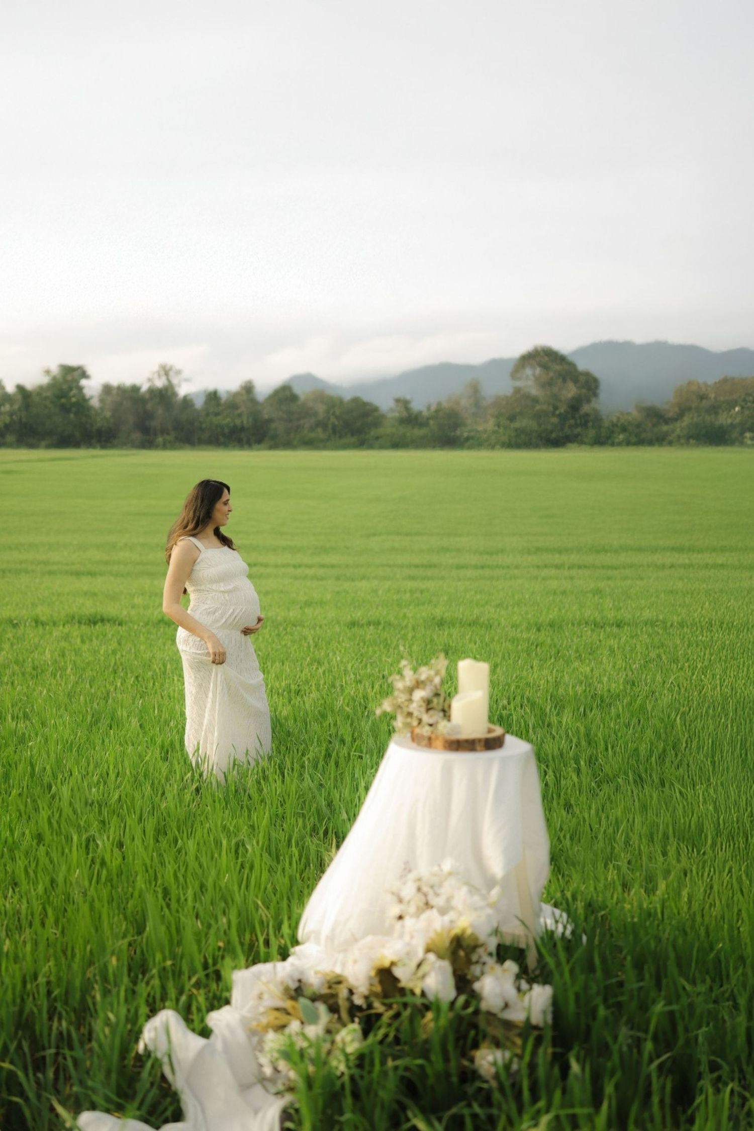 A white-dressed figure stands in a lush green rice field with mountains in the background and a decorated table setting nearby.