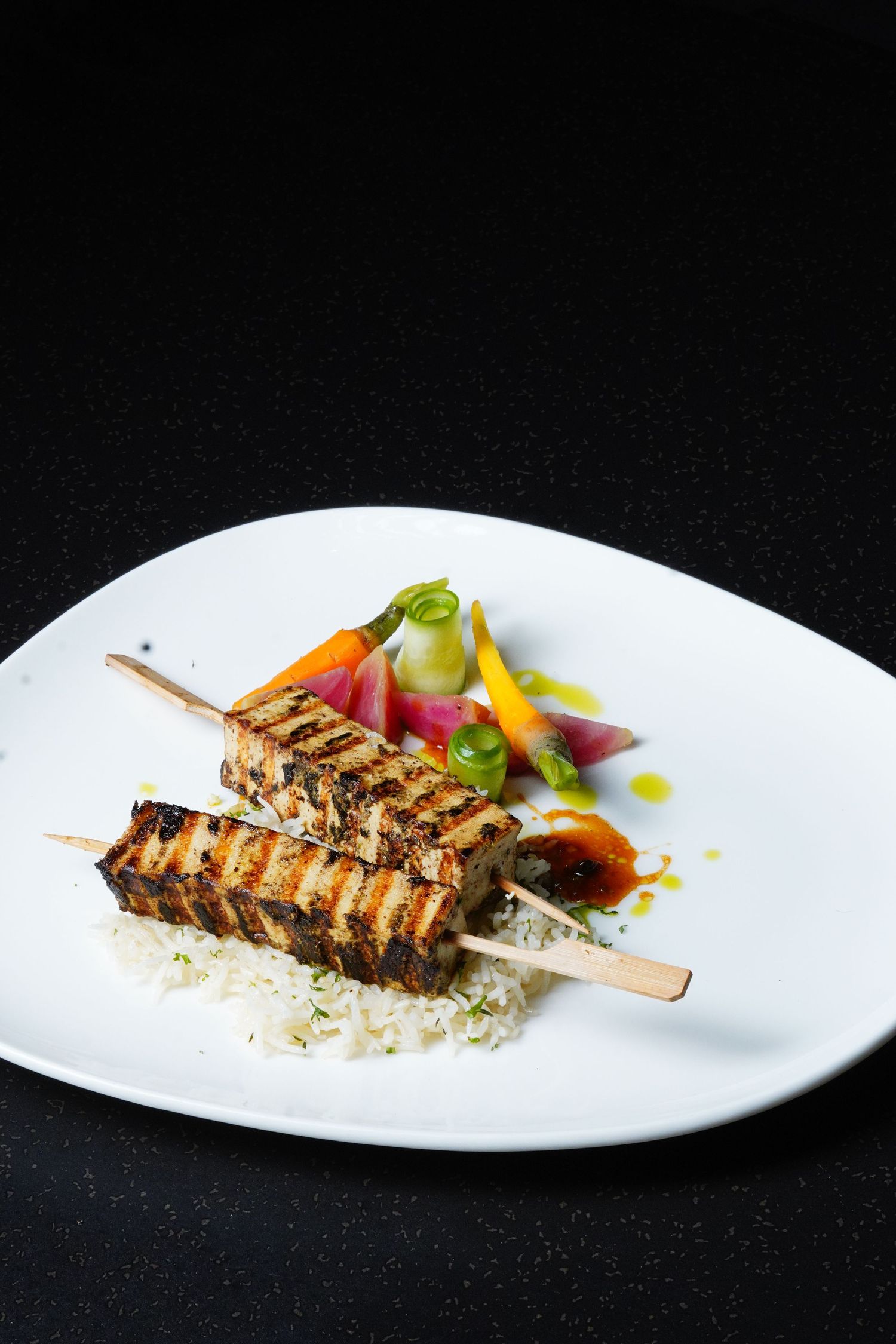 Grilled meat skewers served with colorful vegetables on a white plate against black background.