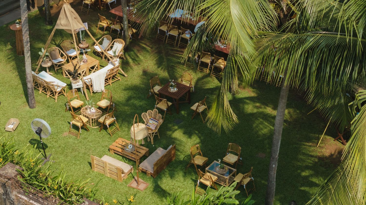 Aerial view of an outdoor tropical restaurant with wooden furniture and tables on lush green grass surrounded by palm trees.