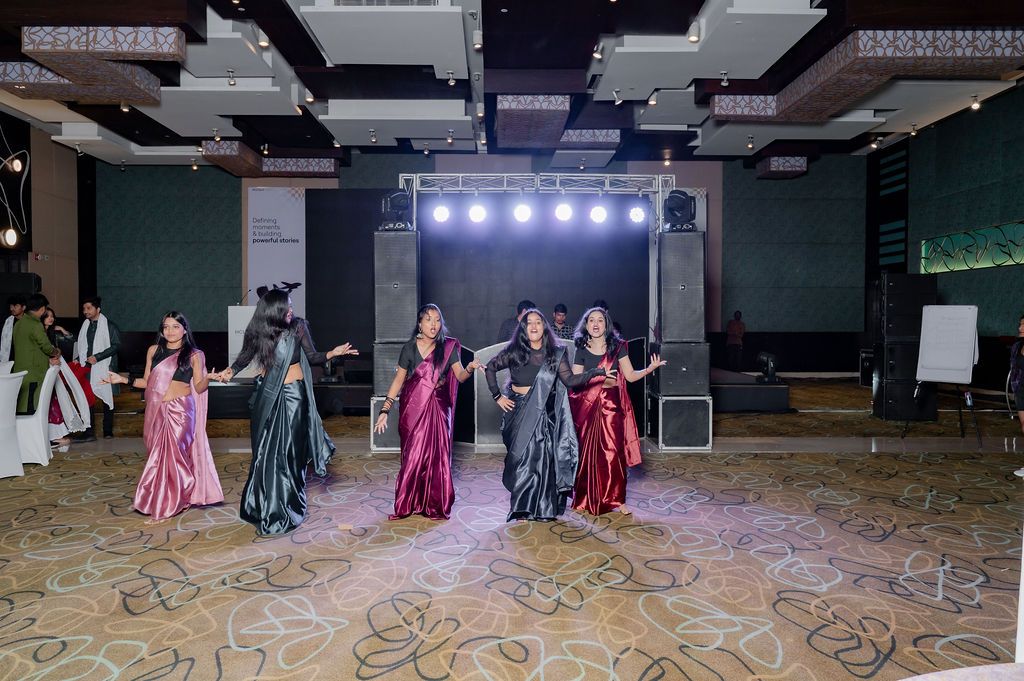 Women in colorful sarees performing a traditional Indian dance on a stage at an indoor event venue.