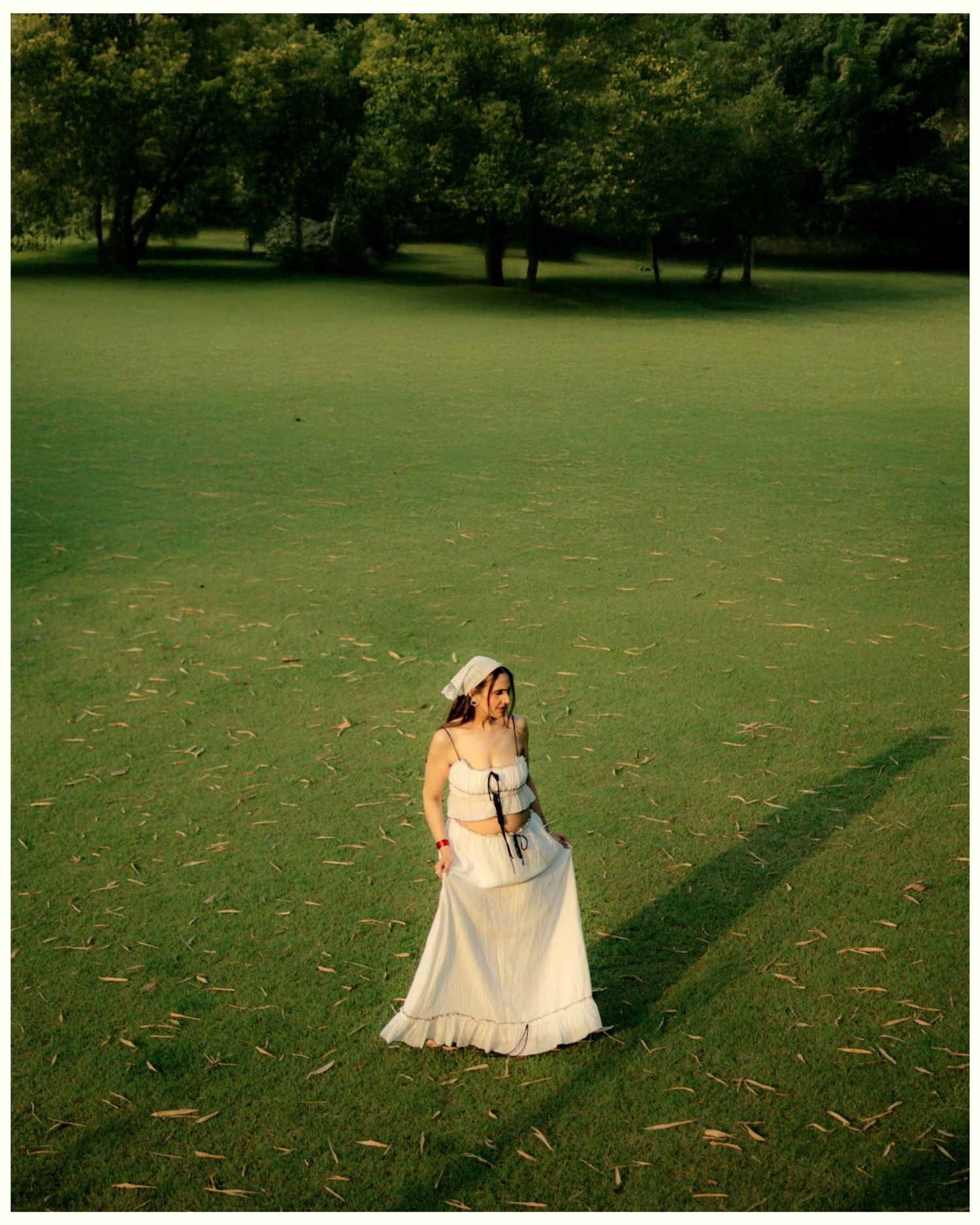 A bride in a white gown and hat stands alone on a lush green lawn surrounded by trees.