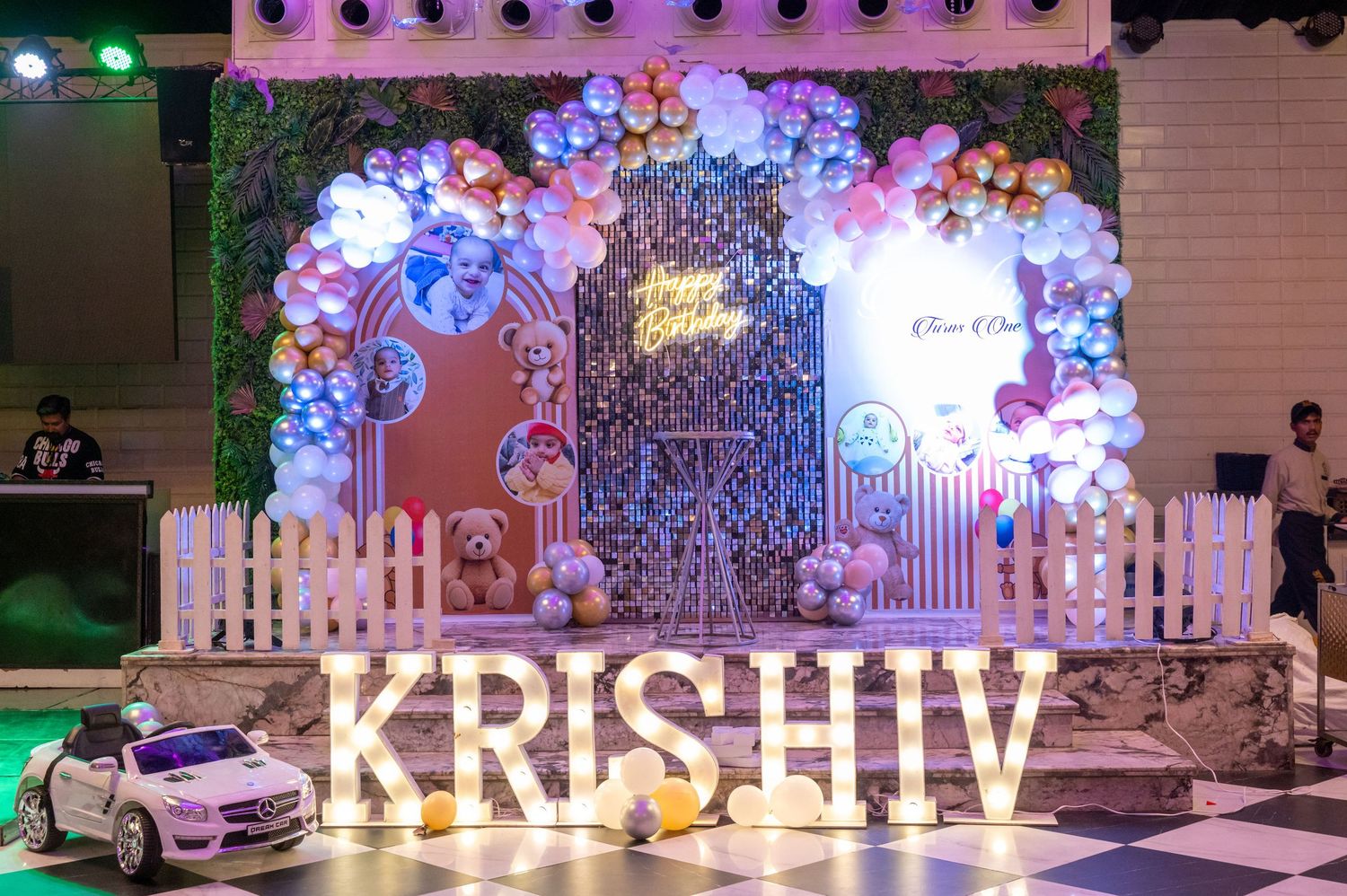 Colorful balloon arch decorates a birthday party backdrop with KRISHIV name display and teddy bear theme decor.