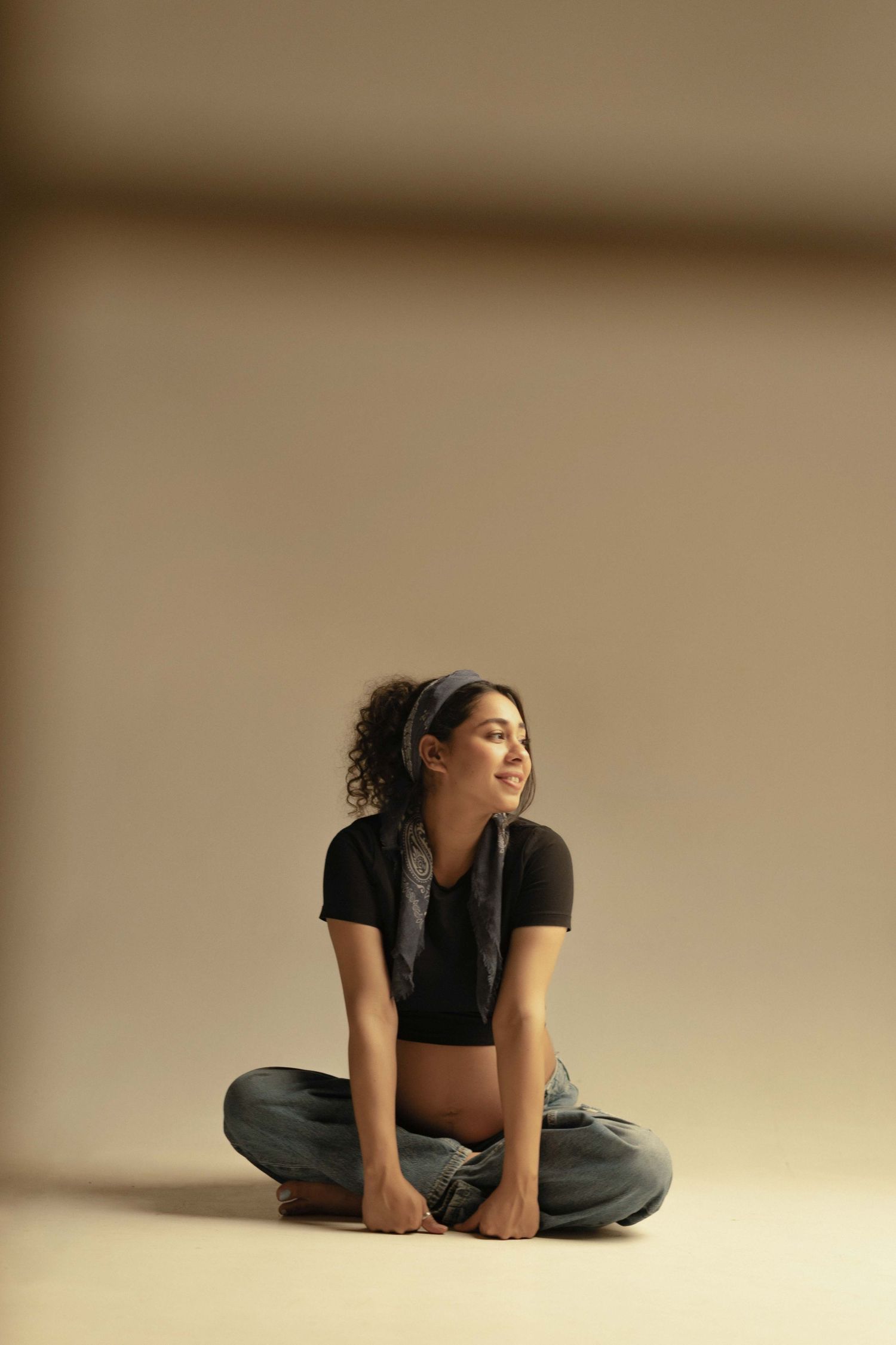 Young woman sitting cross-legged on floor in black crop top and jeans, smiling sideways in neutral beige studio setting.