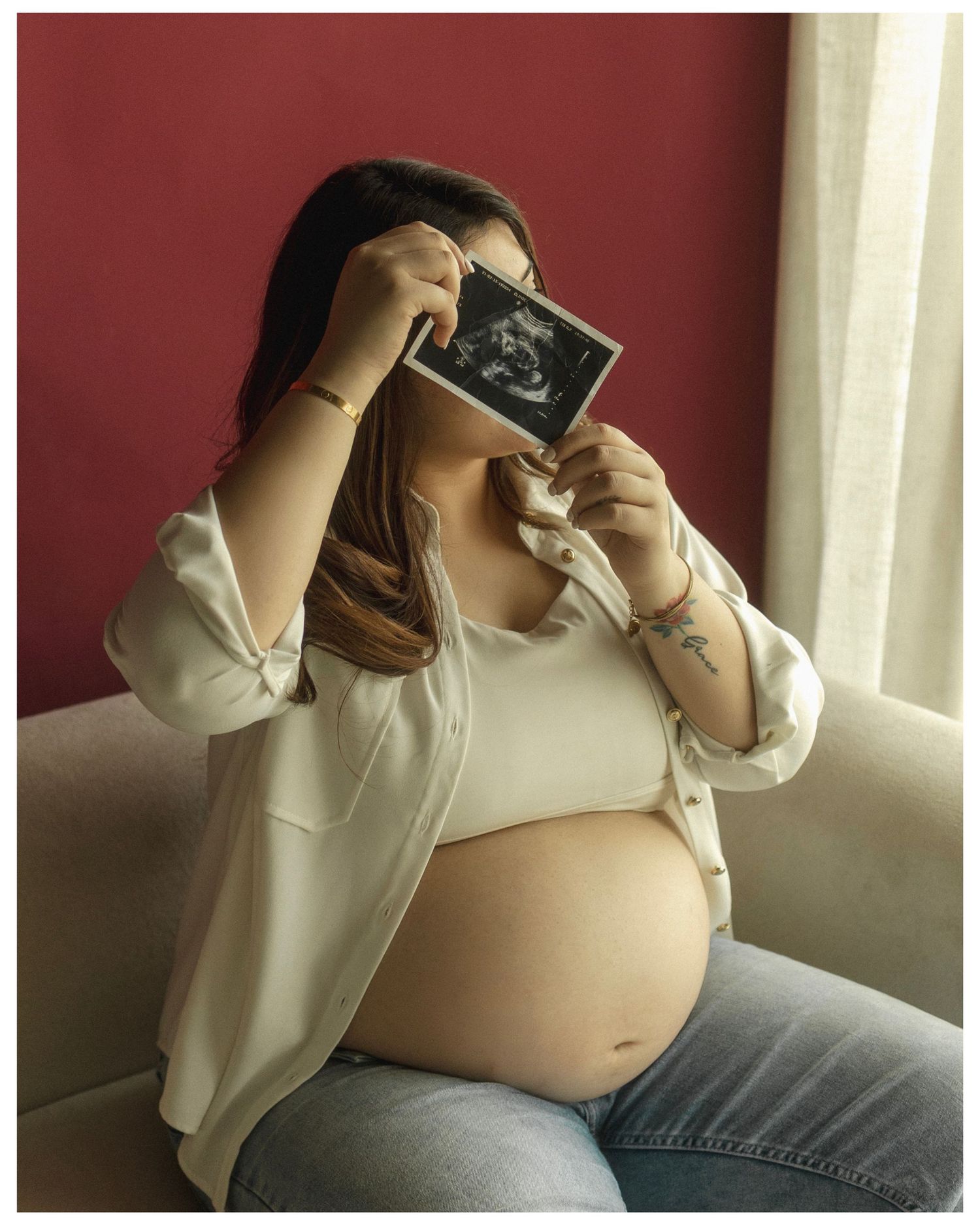 Pregnant woman sitting on a sofa holding an ultrasound photo, wearing a white top and open shirt.
