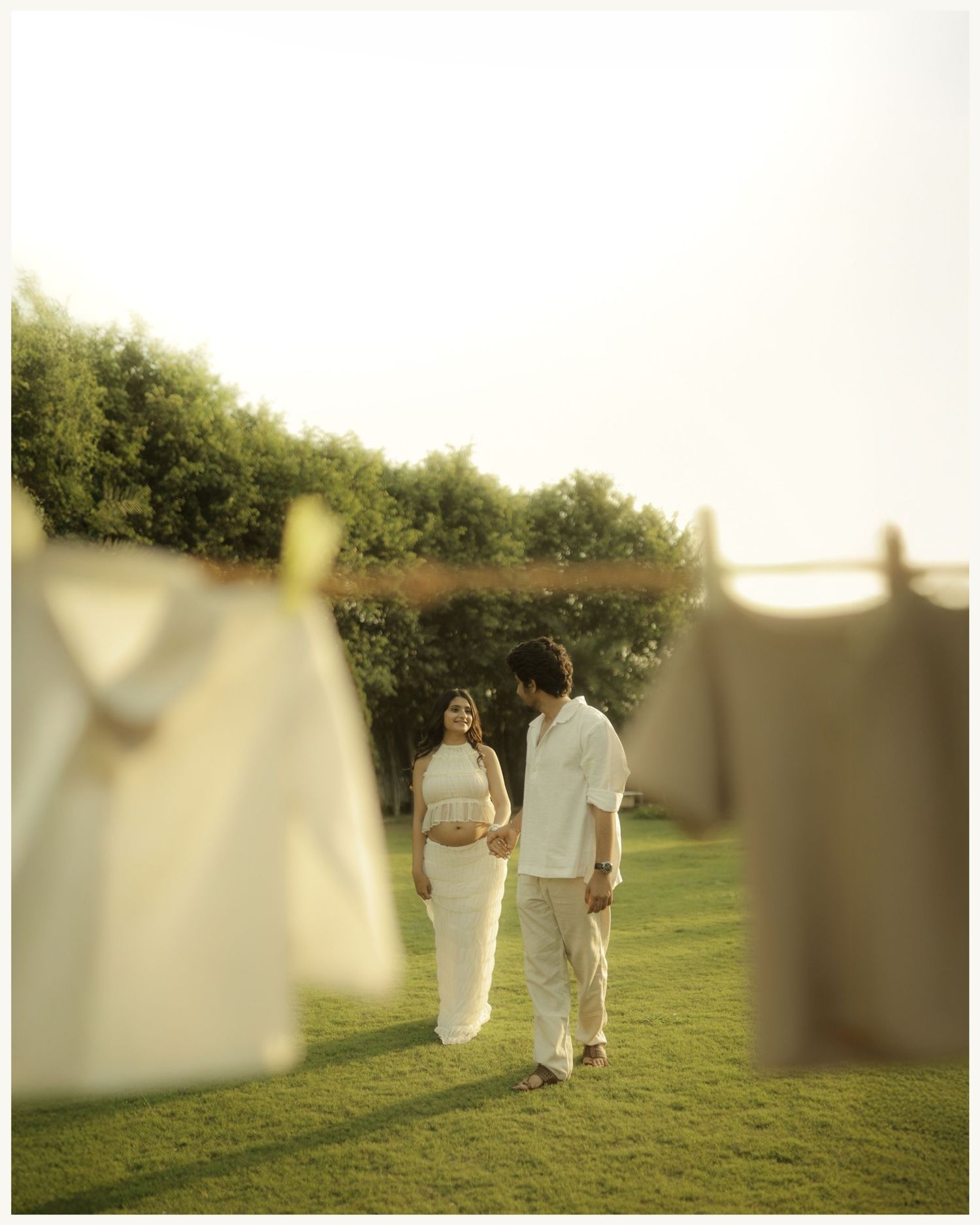 Pregnant couple holding hands on green lawn with white clothes hanging on a clothesline in warm golden light.