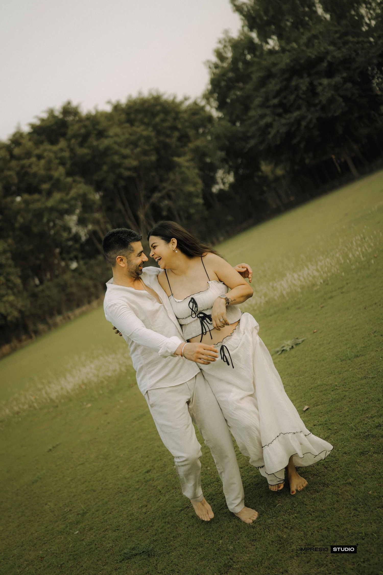 Couple in white outfits dancing barefoot on green grass, sharing a joyful moment outdoors with trees in background.