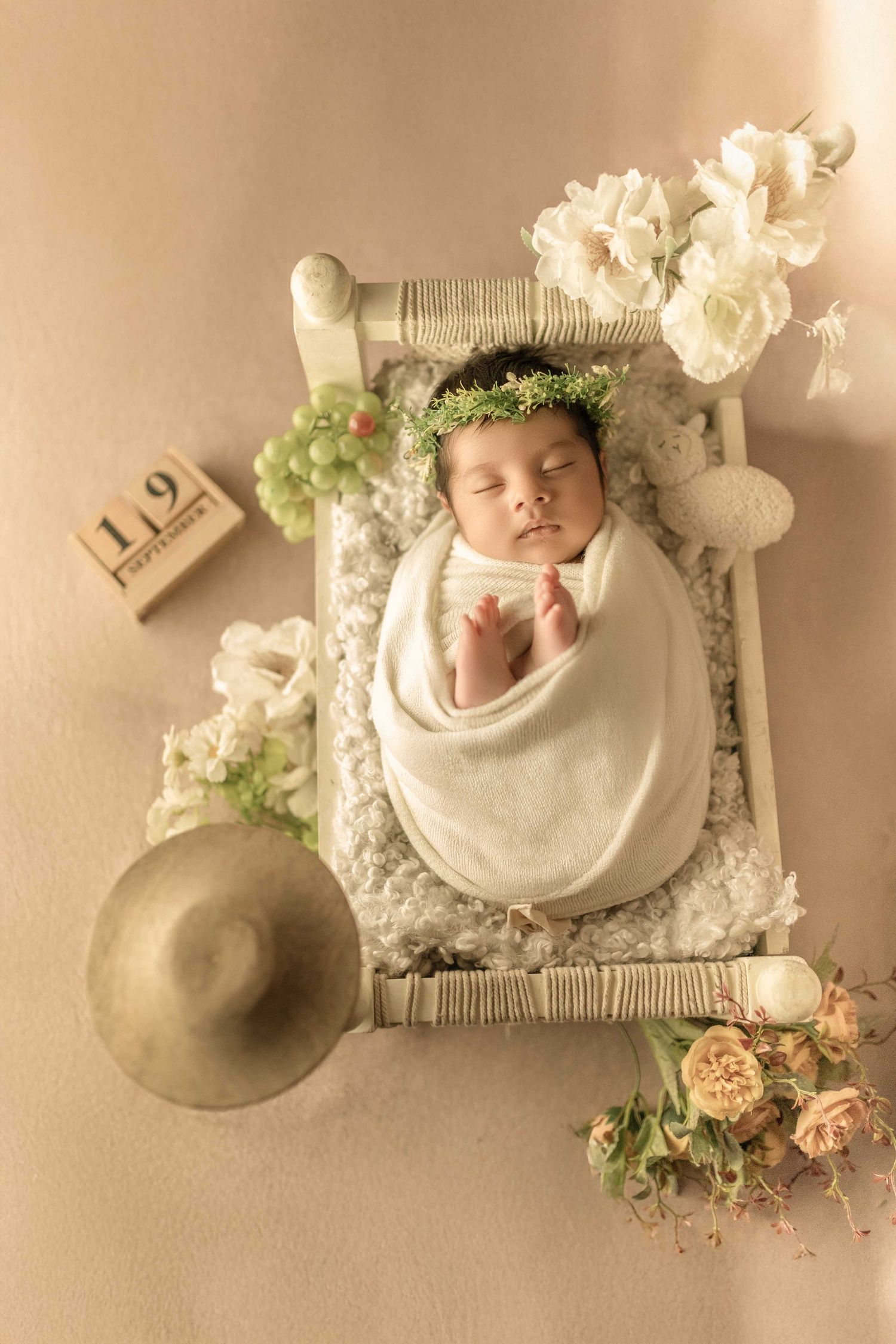 Newborn baby swaddled in white wrap with floral crown sleeping on a tiny decorative bed with flowers and wooden calendar blocks.
