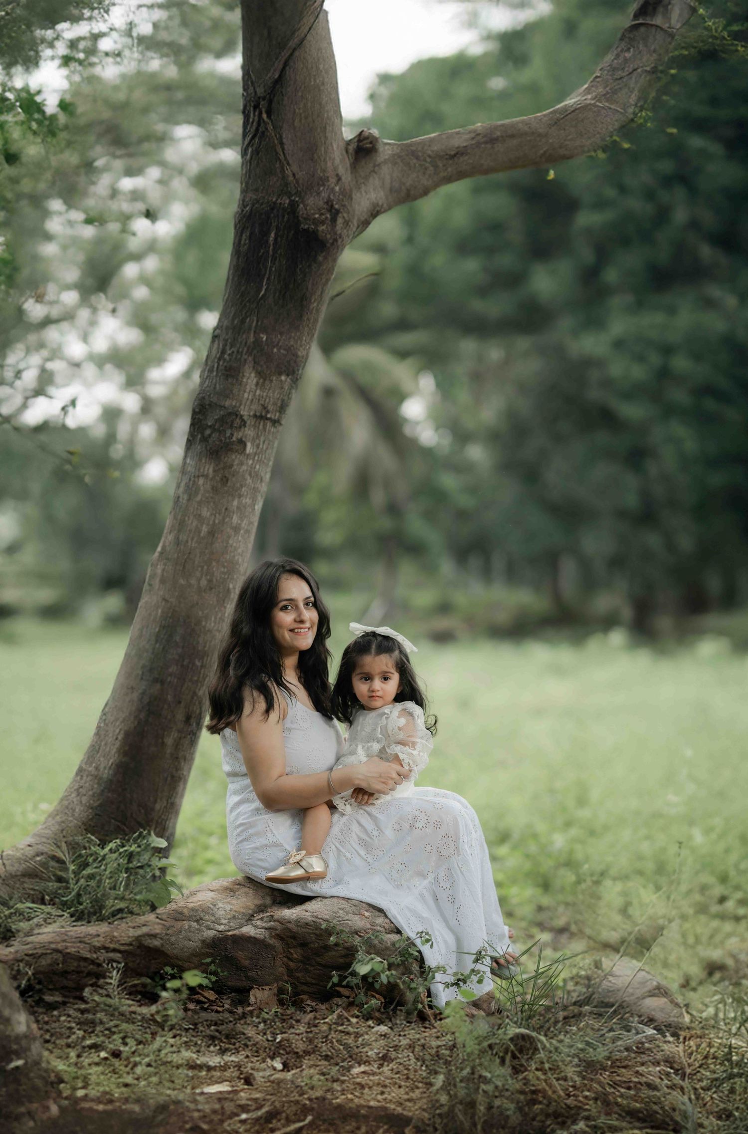 A woman in a white dress sits under a large tree holding a young girl in a lush green park setting.