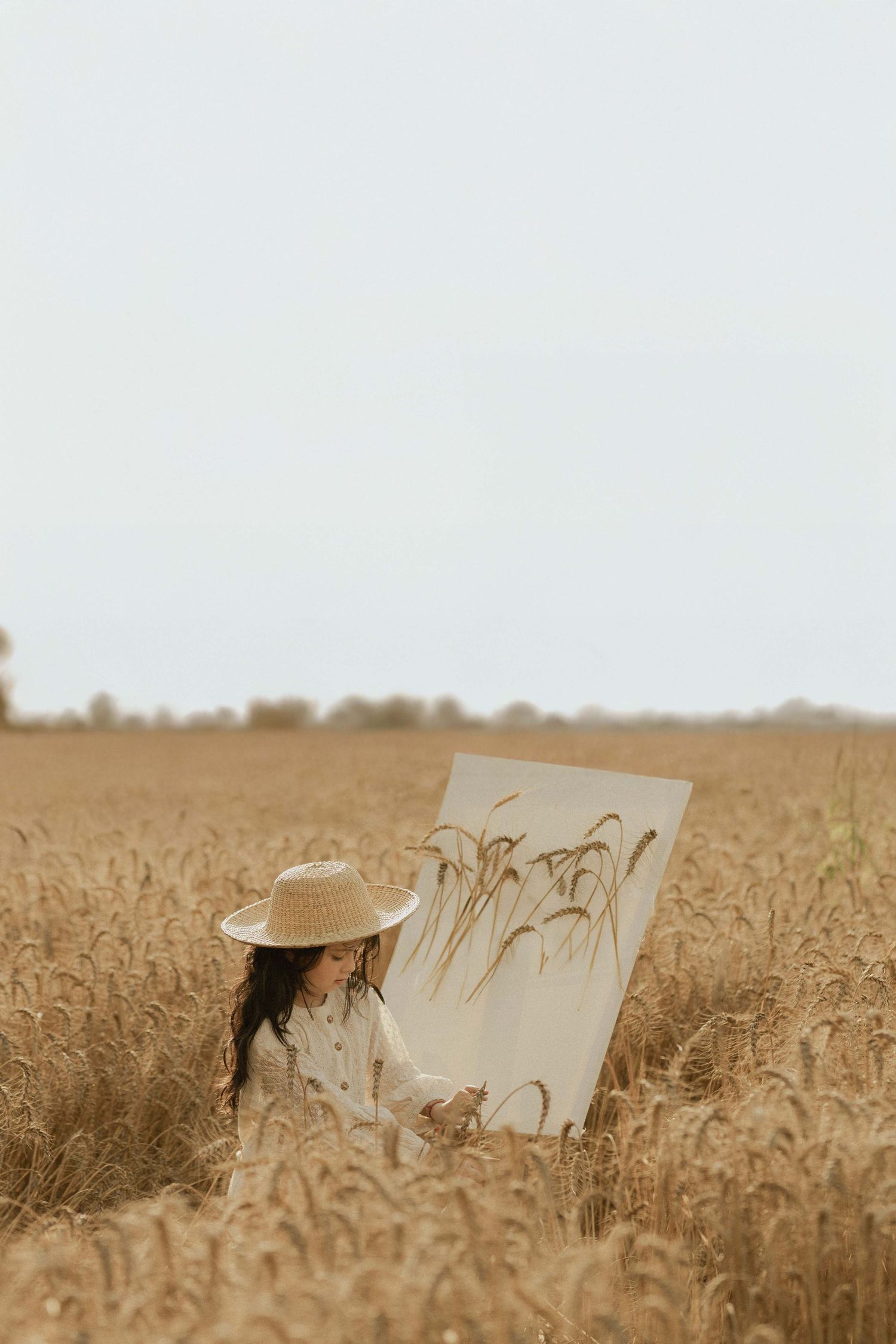 A girl in a straw hat paints wheat stalks on canvas while sitting in a golden wheat field.