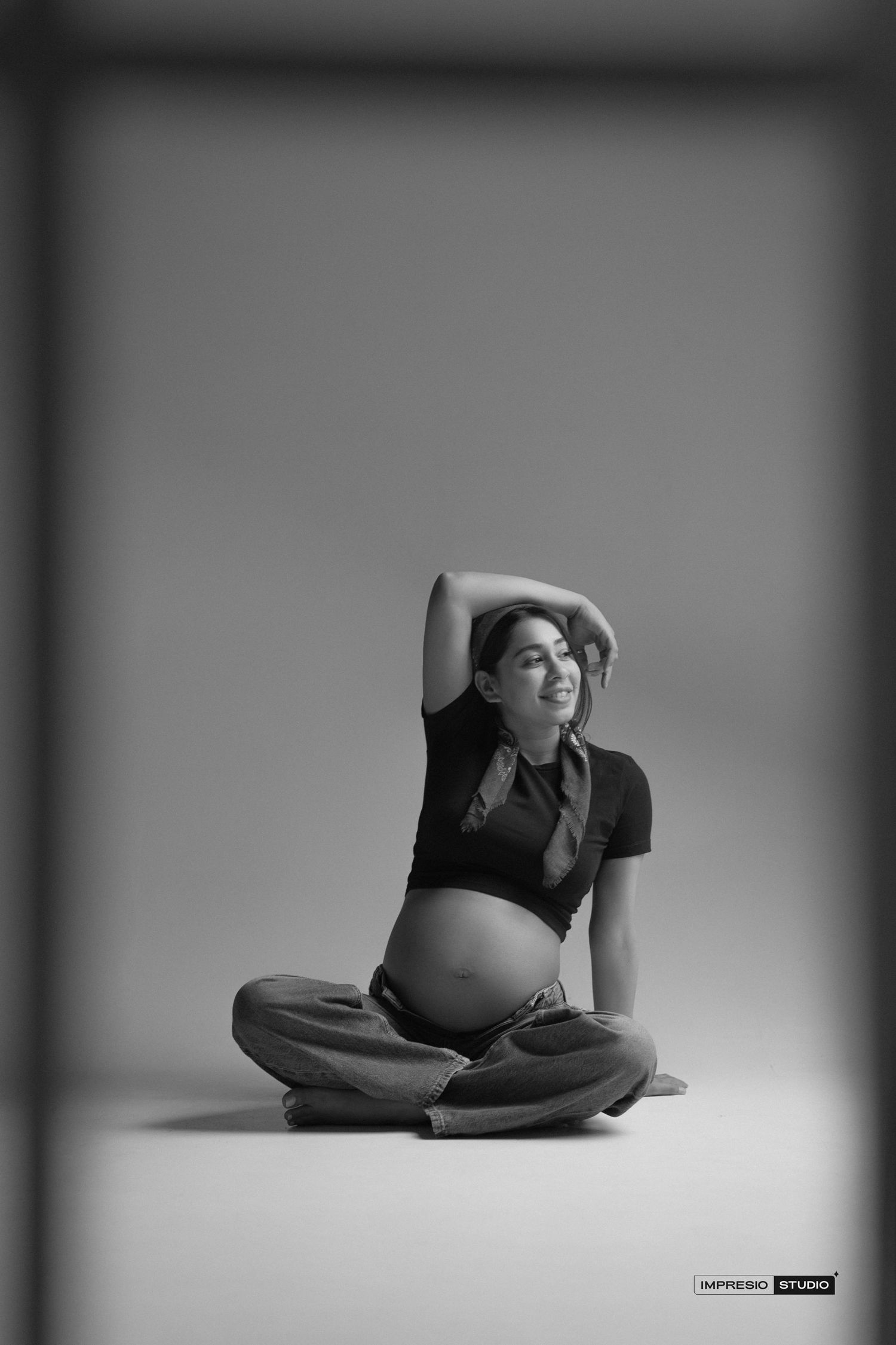 Black and white maternity photo of a pregnant woman sitting cross-legged with arm raised, showcasing her baby bump.