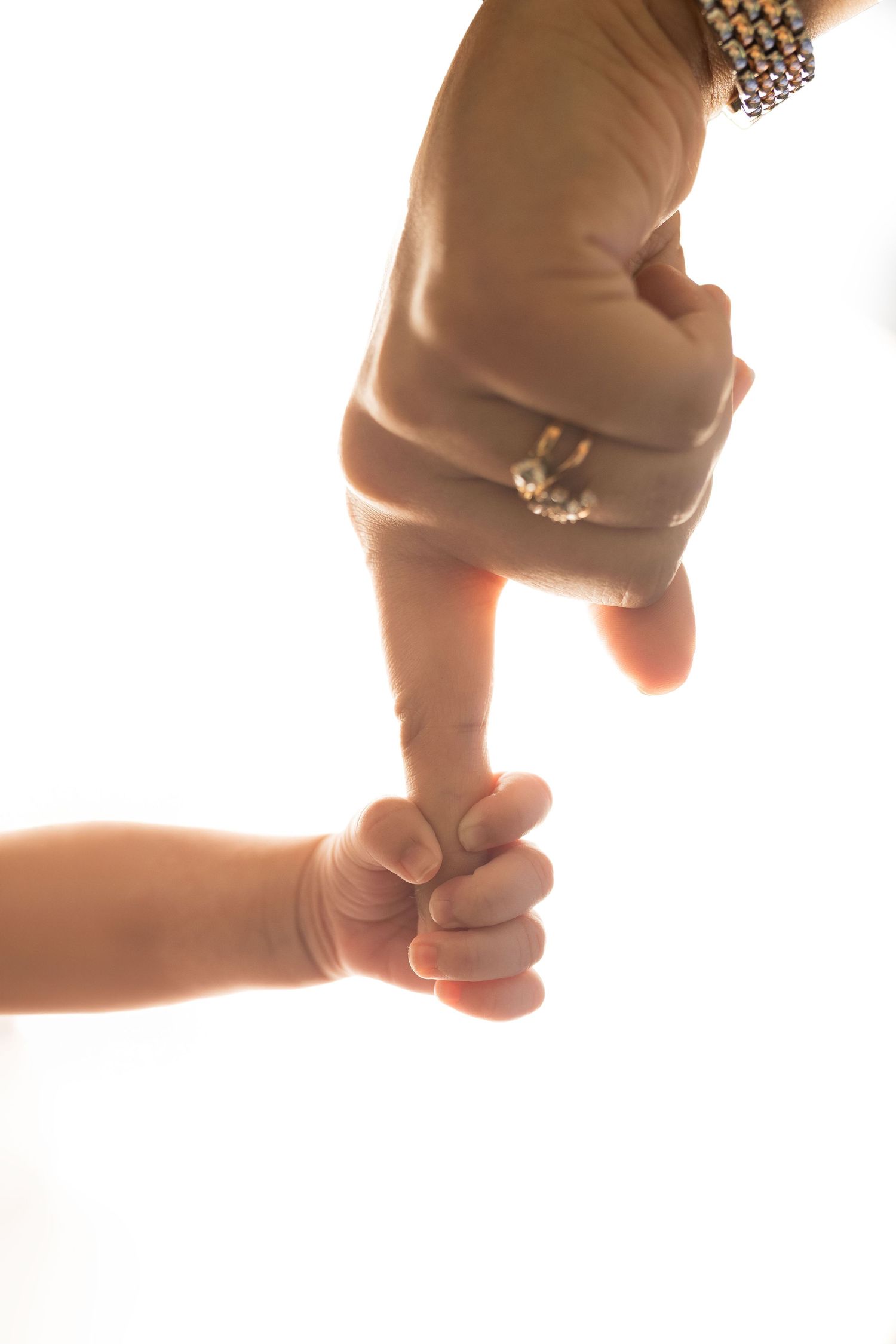 A baby's tiny hand gripping an adult's finger, wearing a ring and bracelet, against a bright white background.