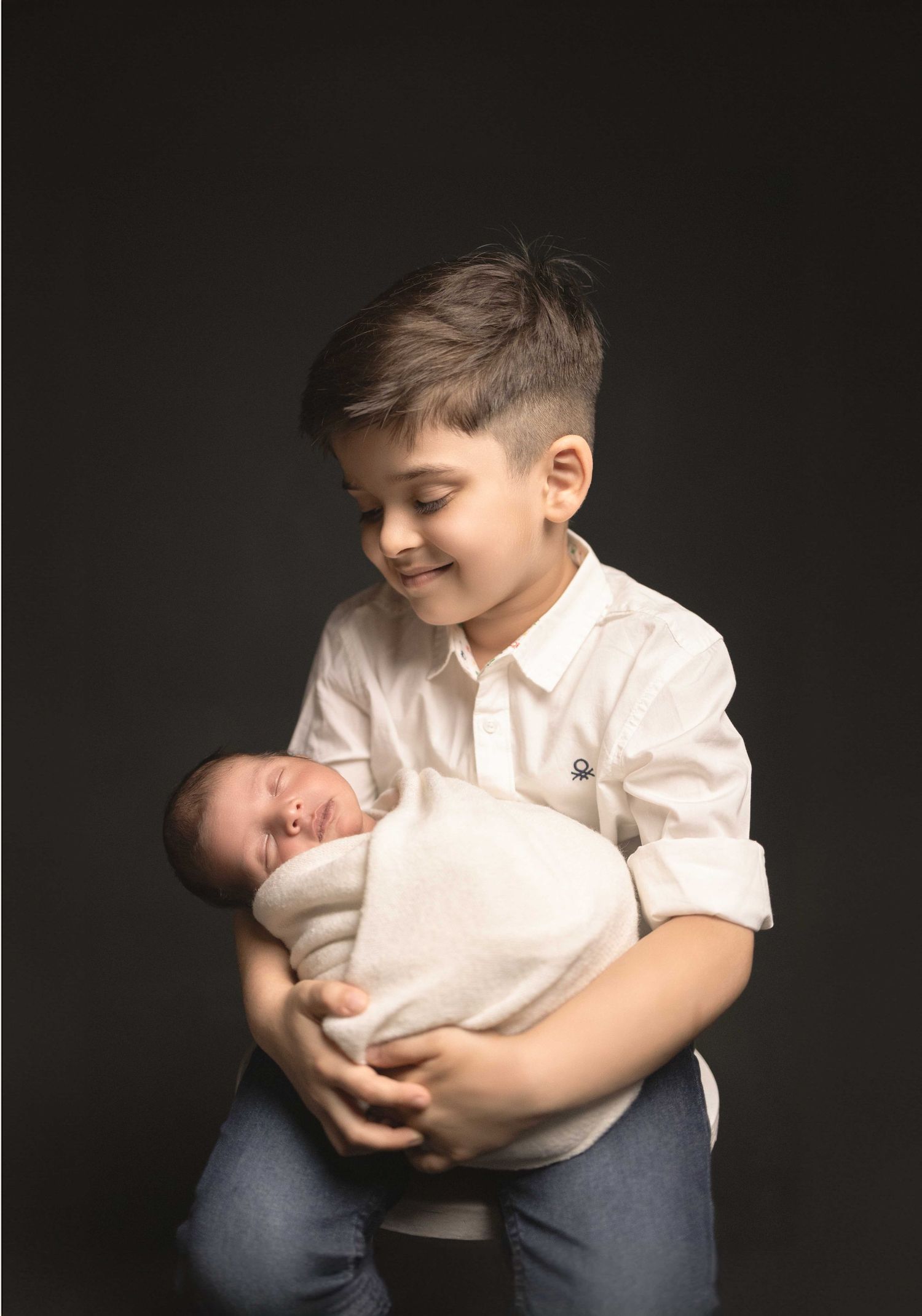A young boy smiles warmly while carefully holding a swaddled newborn baby in a studio portrait.