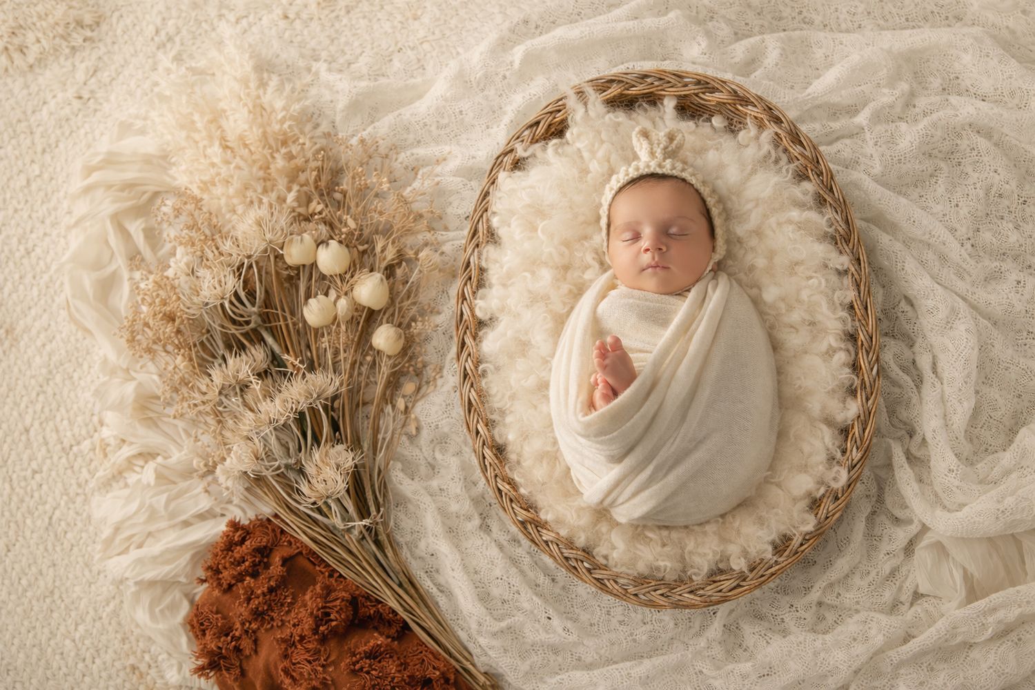 Newborn baby swaddled in white wrap sleeping in wicker basket with cream fur and dried floral arrangement.
