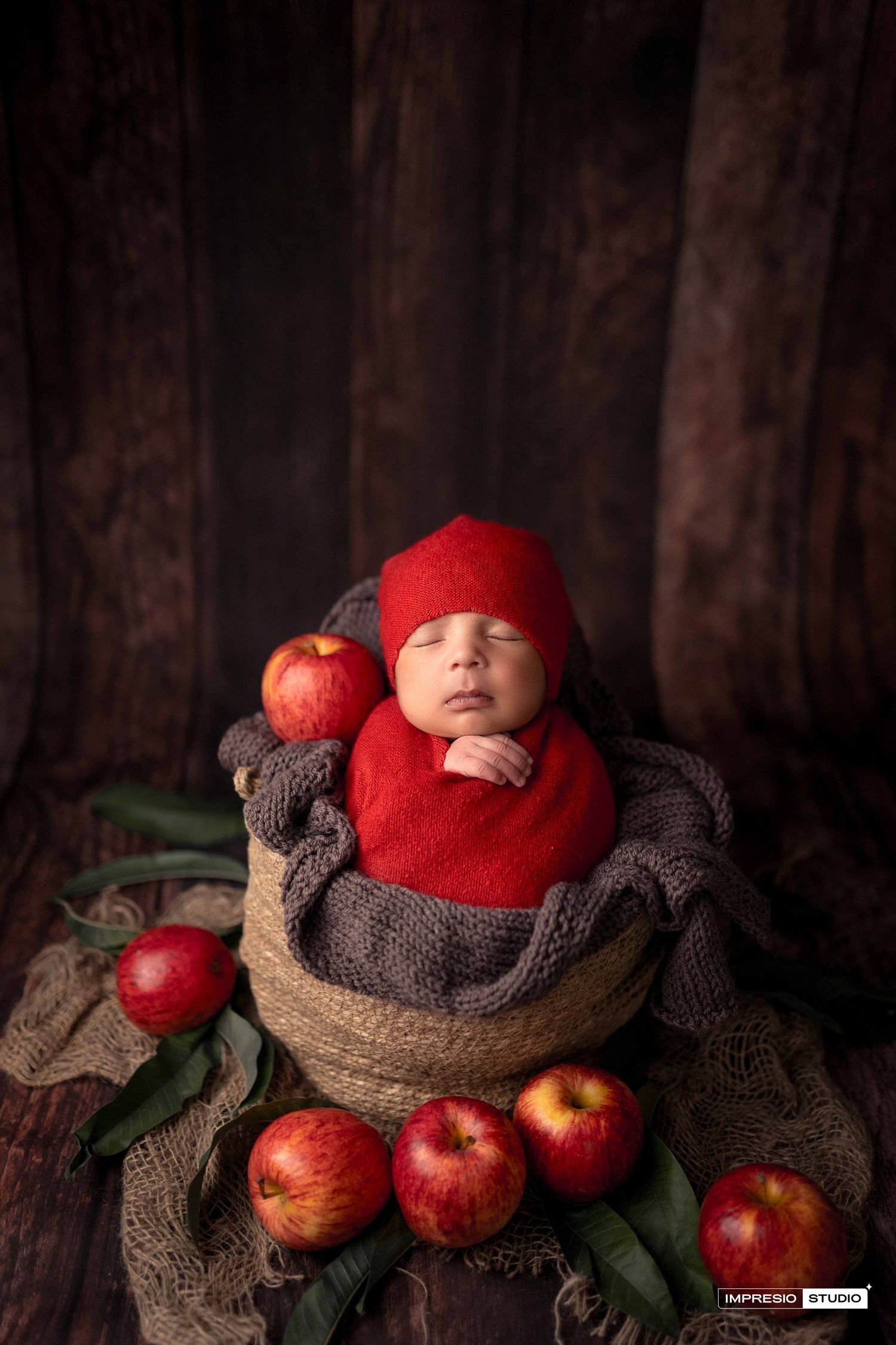 Newborn baby wrapped in red swaddled in a rustic basket surrounded by fresh red apples in a cozy studio portrait.