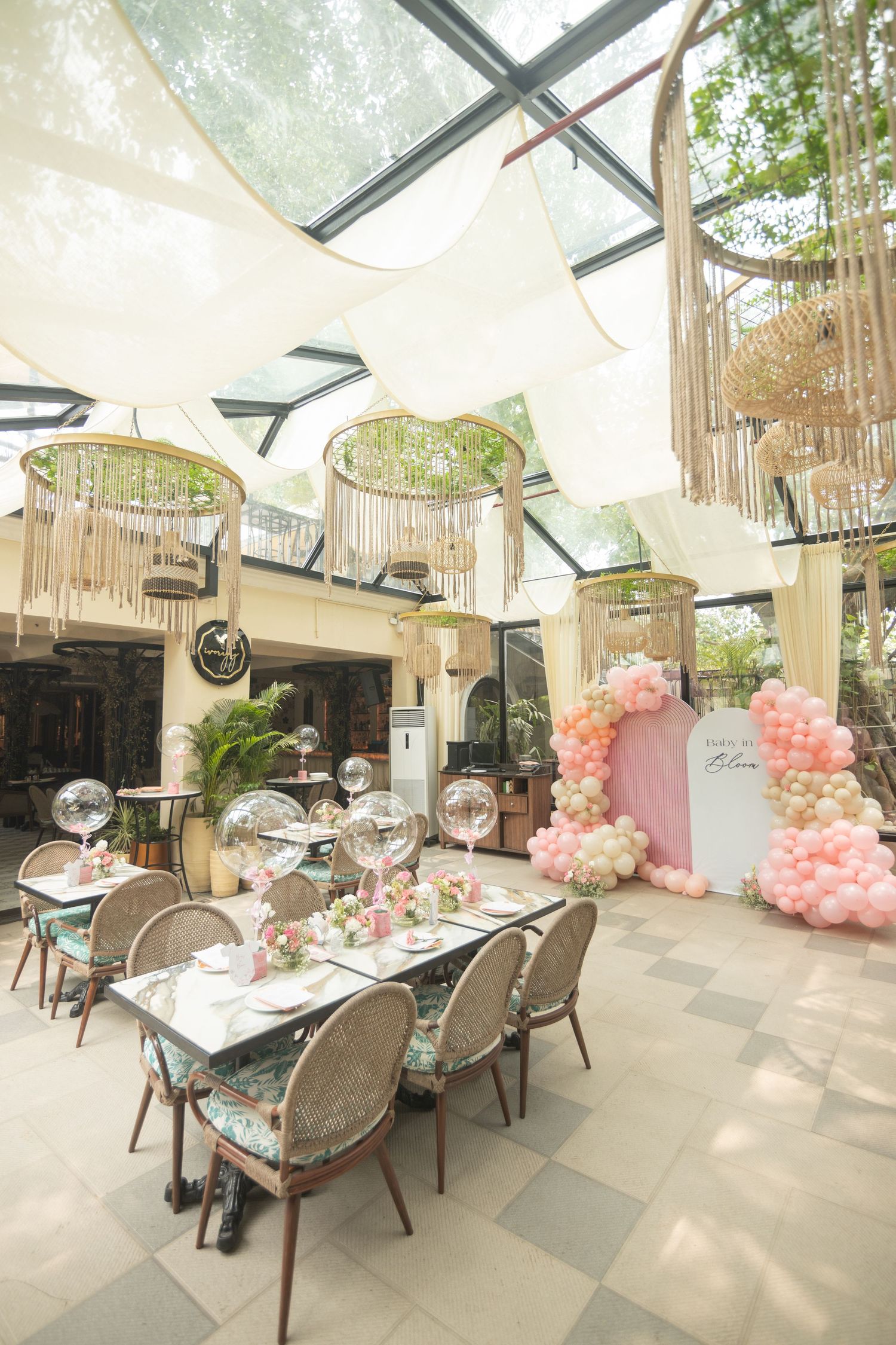 Elegant event space with pink balloon arch, rattan chairs, floral table settings, and a glass ceiling draped with white canopies.
