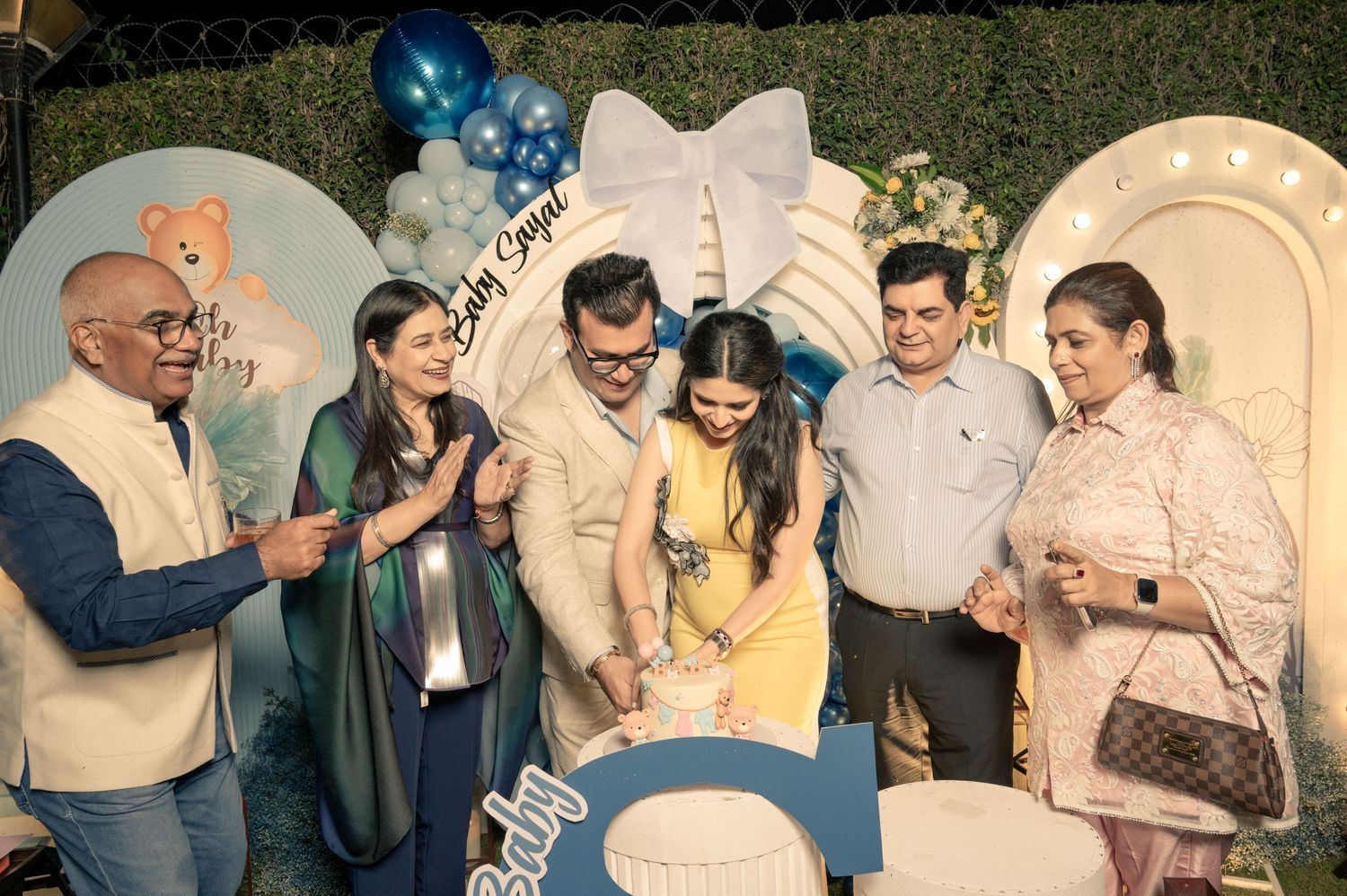 Family and friends gather around a cake at a blue-themed baby shower celebration with balloons and decorations.