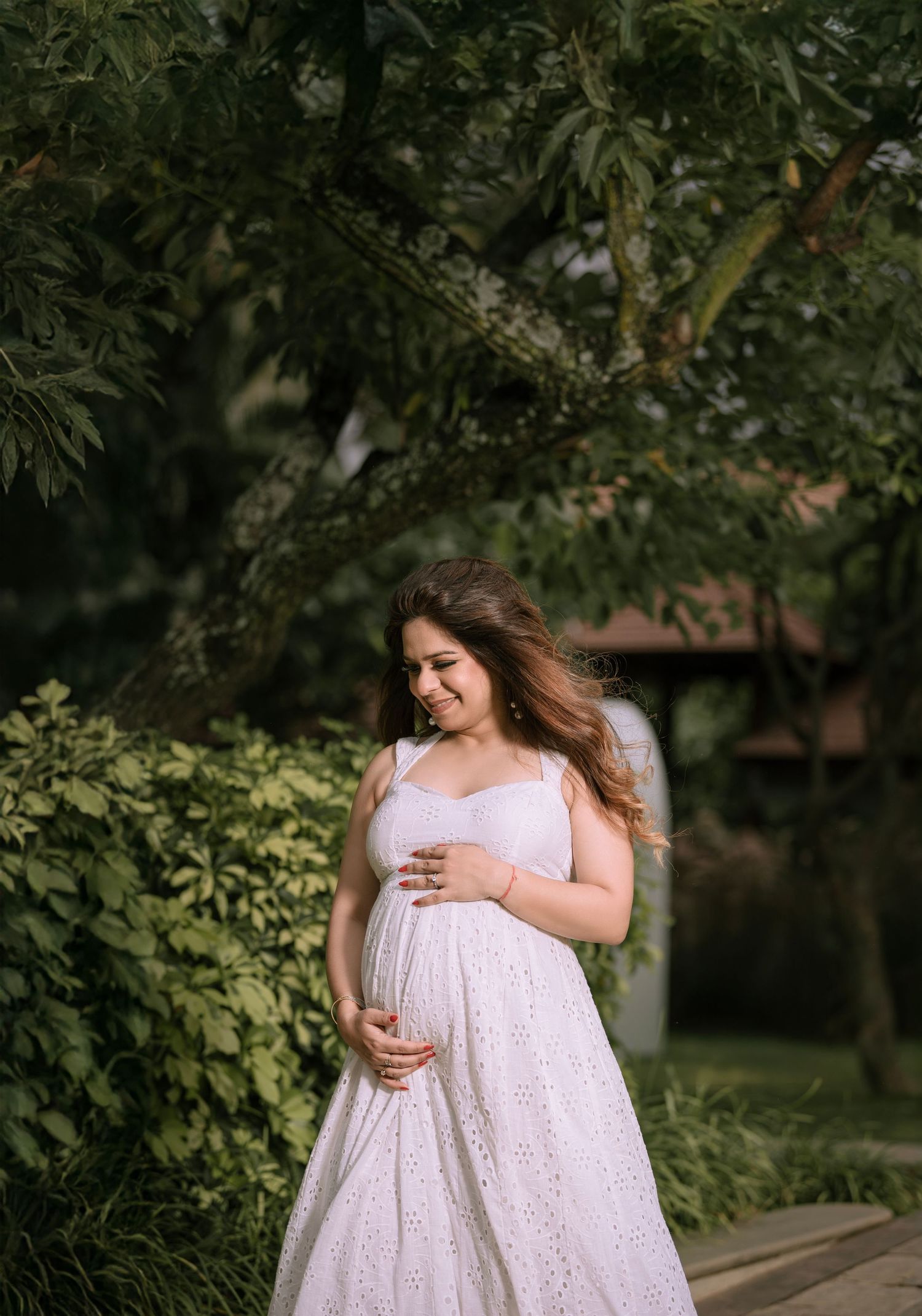 Pregnant woman in white eyelet dress cradling baby bump outdoors among lush green trees.