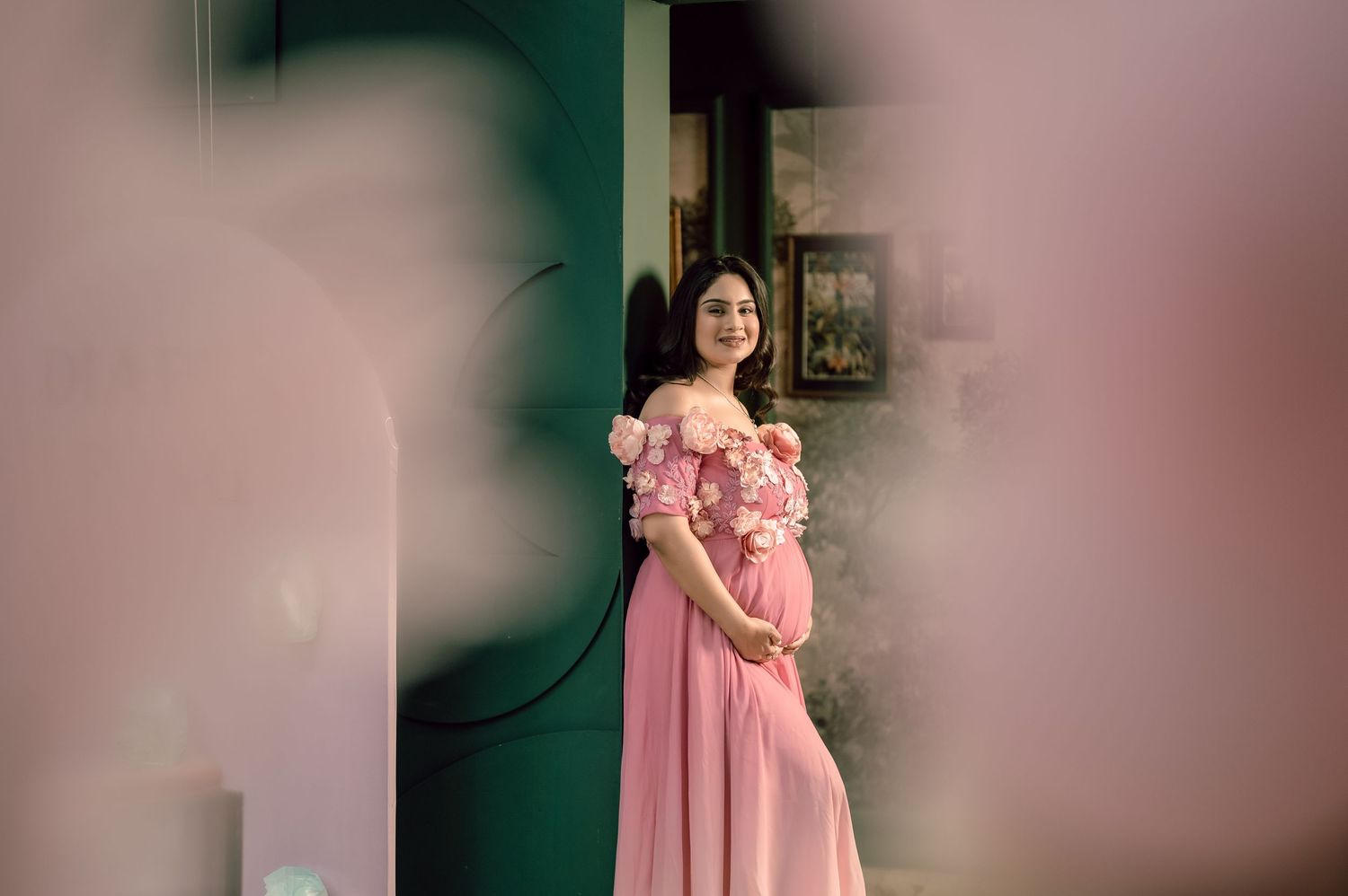 Pregnant woman in pink off-shoulder floral gown posing elegantly outdoors near a teal door.