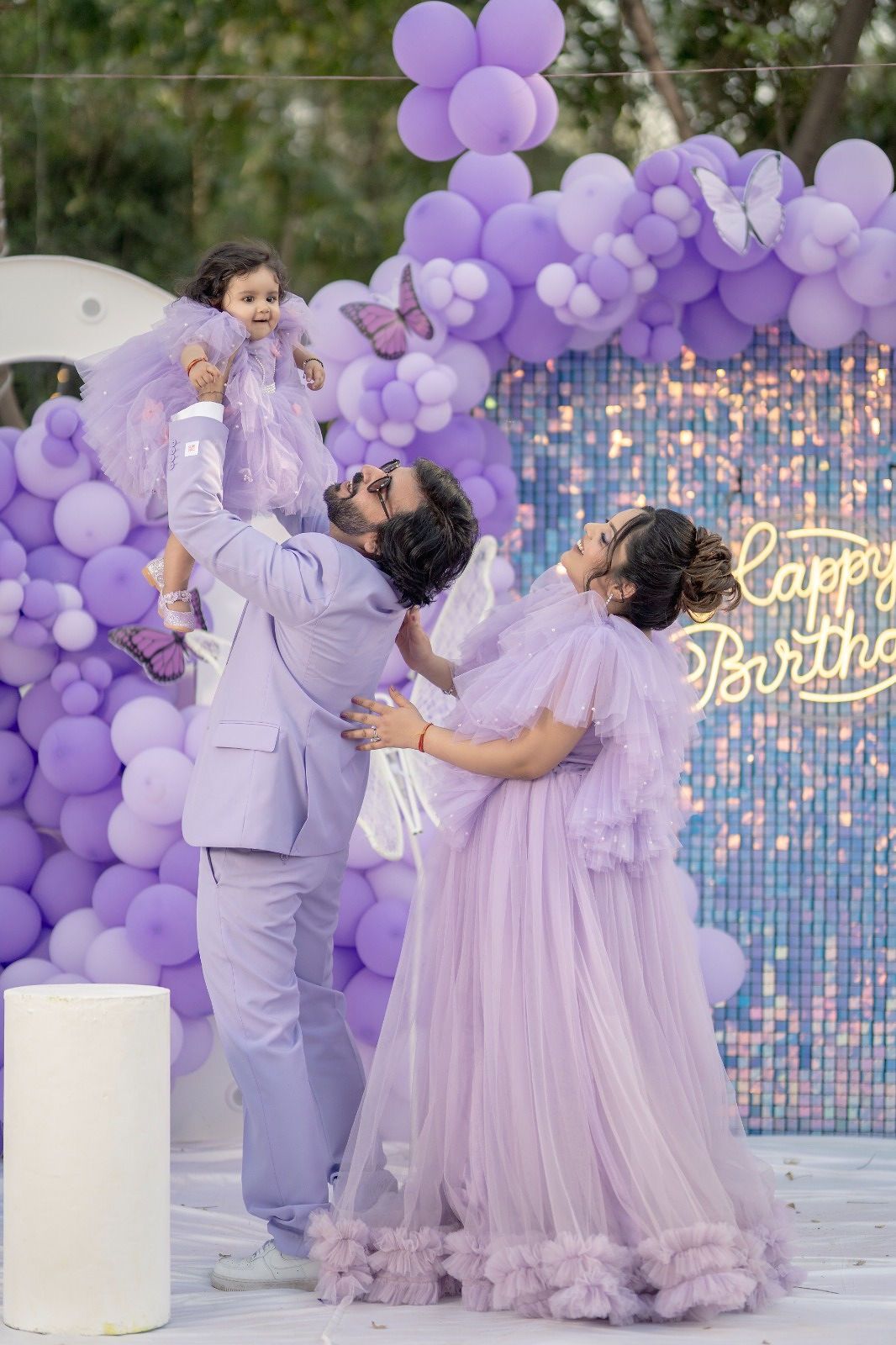 Family in matching lavender outfits celebrating a birthday with purple balloon arch and Happy Birthday backdrop.