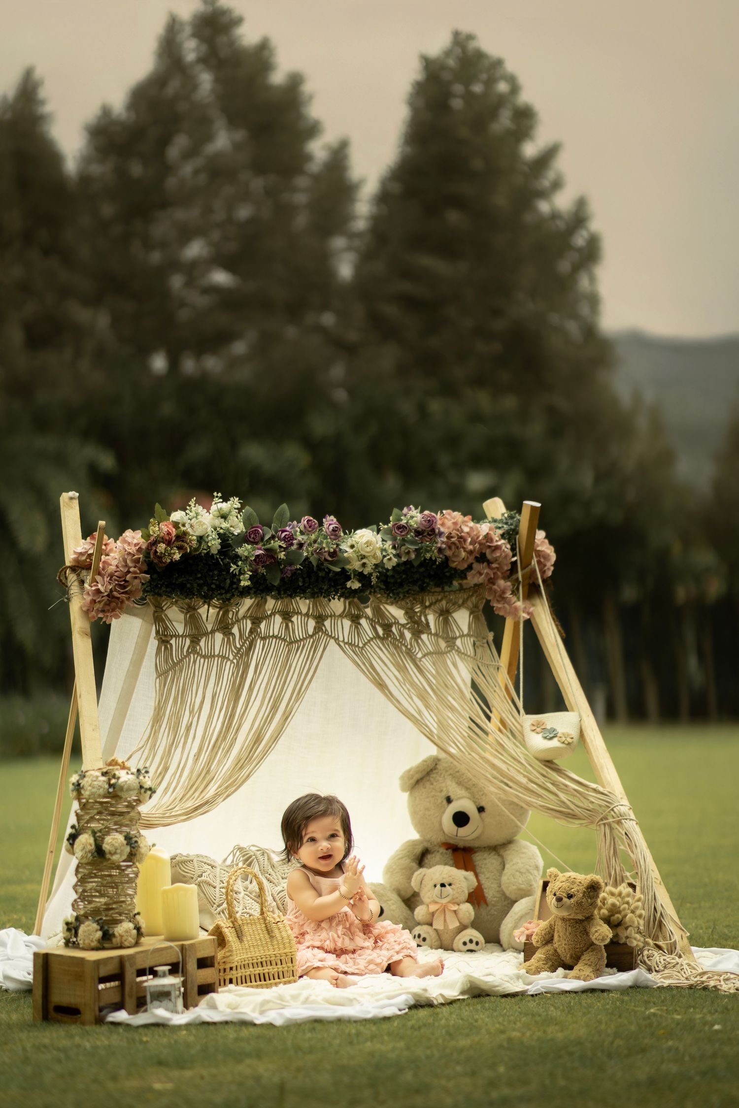 Baby girl in pink dress sits smiling under floral boho canopy with teddy bears on grass outdoors.