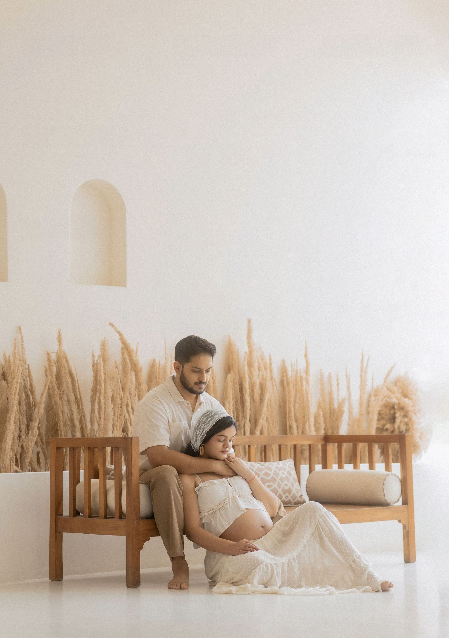 Couple sharing a tender maternity photo shoot moment on a wooden bench with pampas grass backdrop.