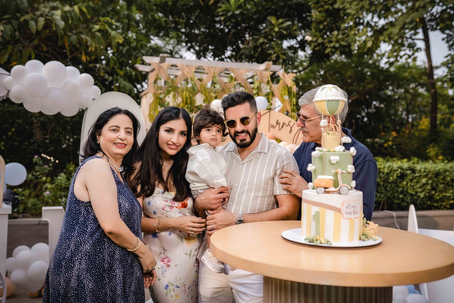 Family celebrating a birthday outdoors with a decorated cake, balloons, and a floral backdrop at a festive party.