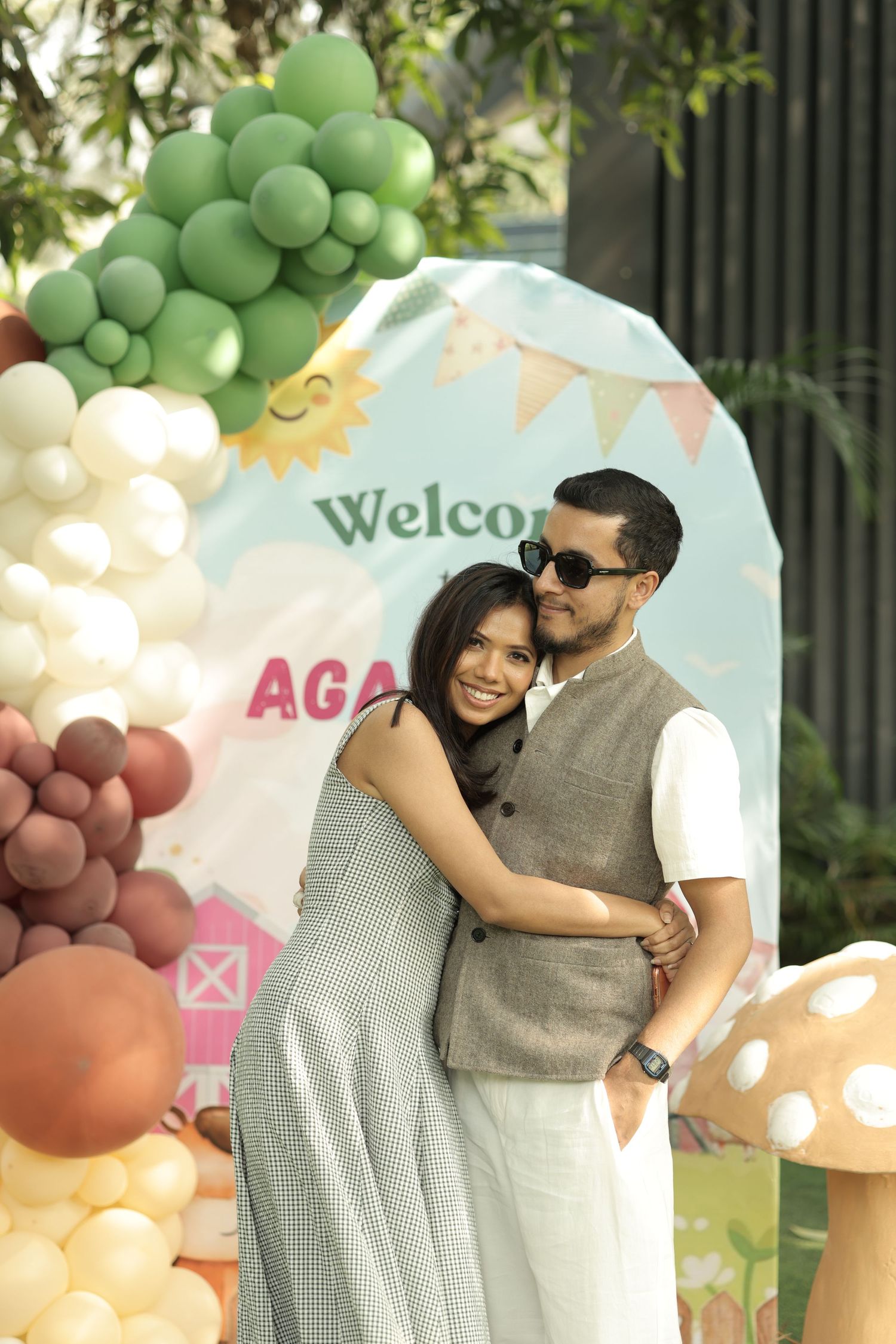 Smiling couple embraces at a colorful balloon arch party backdrop with Welcome sign and festive decorations.