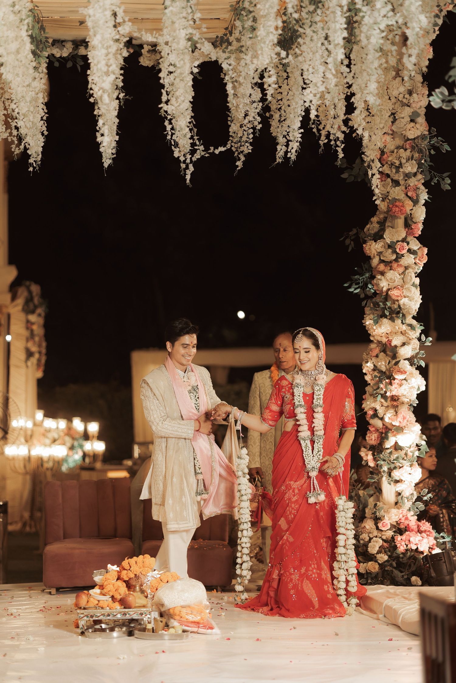 Bride in red saree and groom in beige sherwani perform Hindu wedding rituals under floral mandap at night.