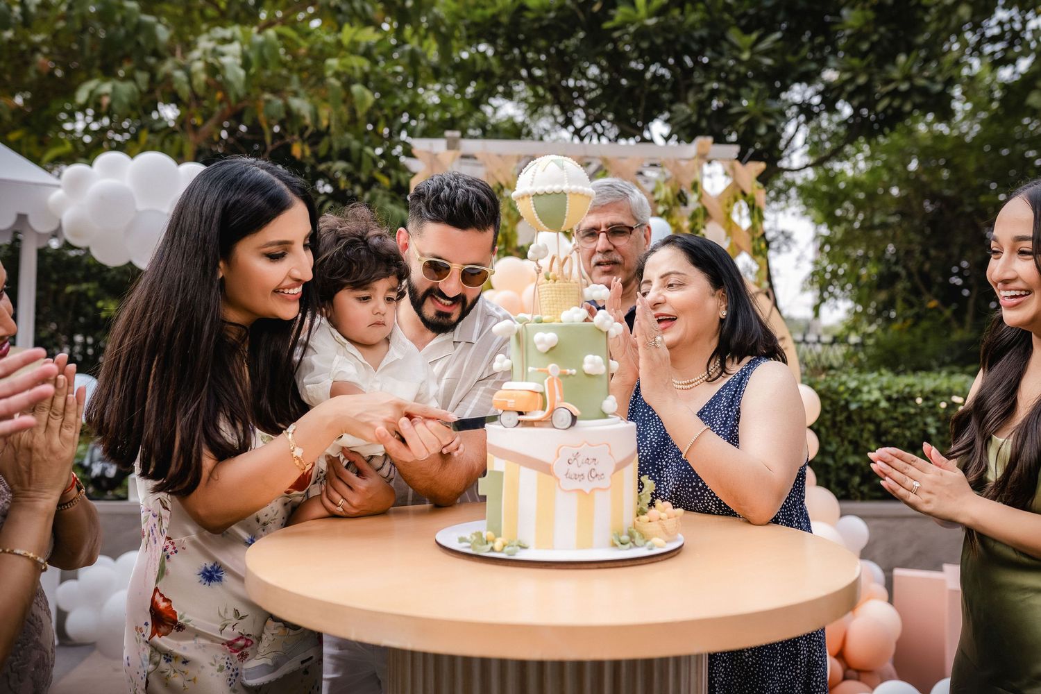 Family gathered around a decorated birthday cake outdoors, celebrating together with joy and smiles.