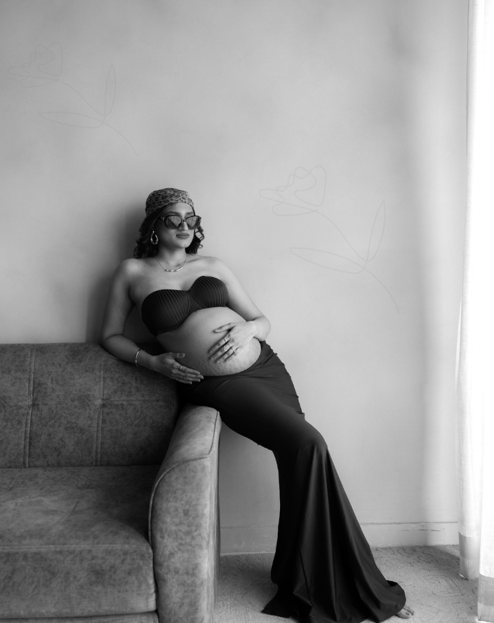 Black and white maternity photo of a pregnant woman in a strapless gown sitting on a sofa, cradling her baby bump.