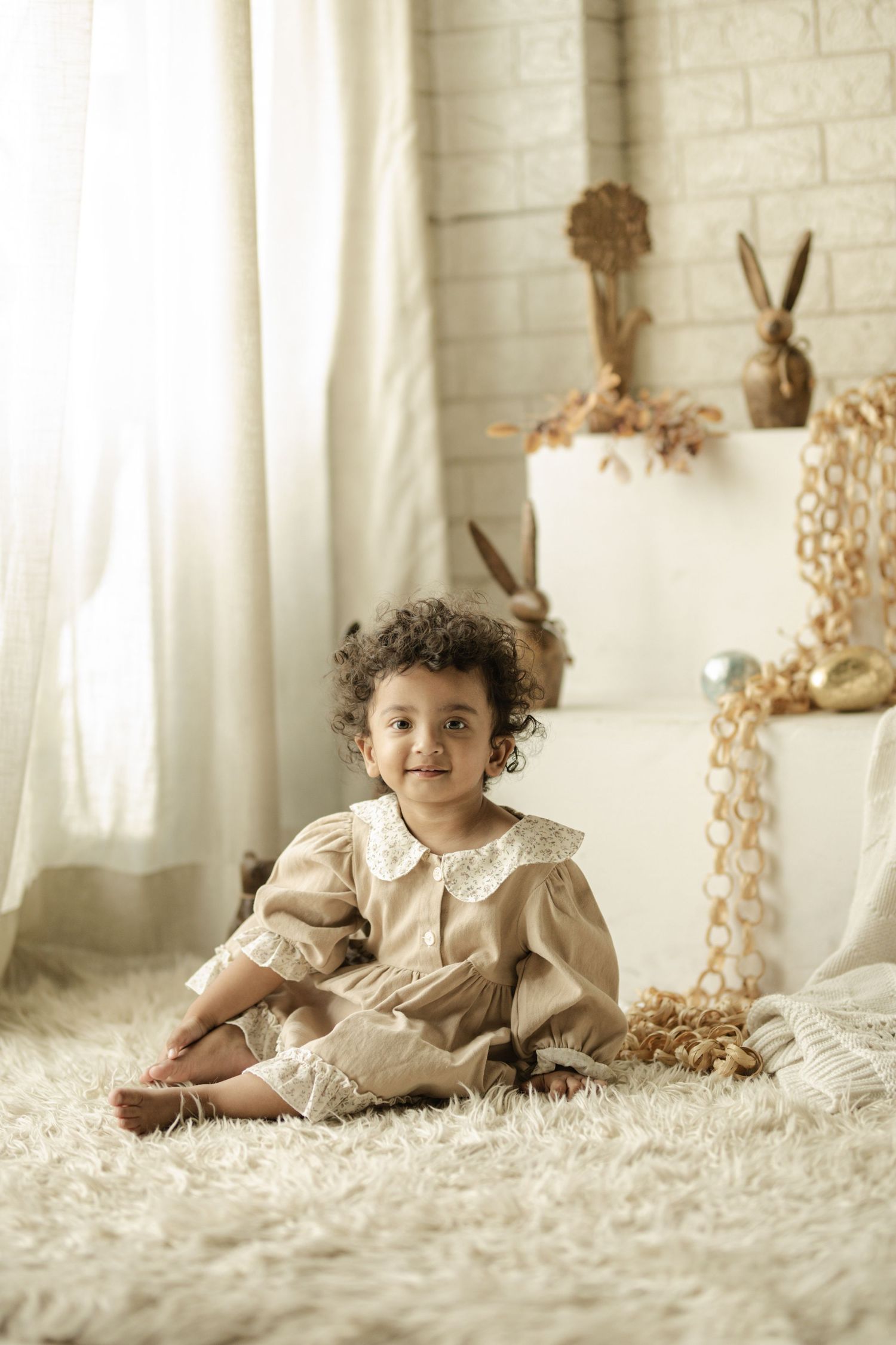 Smiling toddler girl in beige lace dress sitting on white fluffy rug in bright room with Easter decorations.