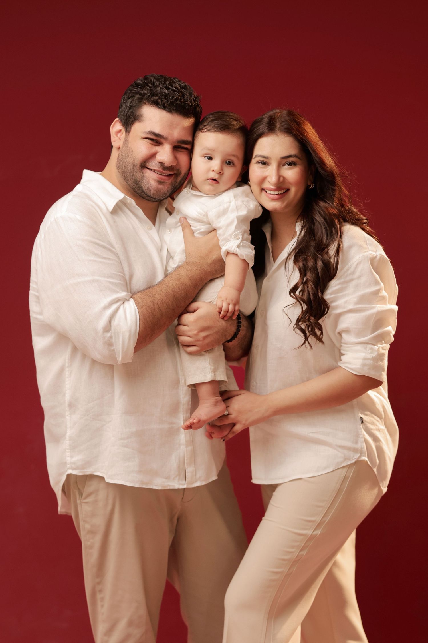 Happy family portrait with parents in white shirts holding their baby against a red background.