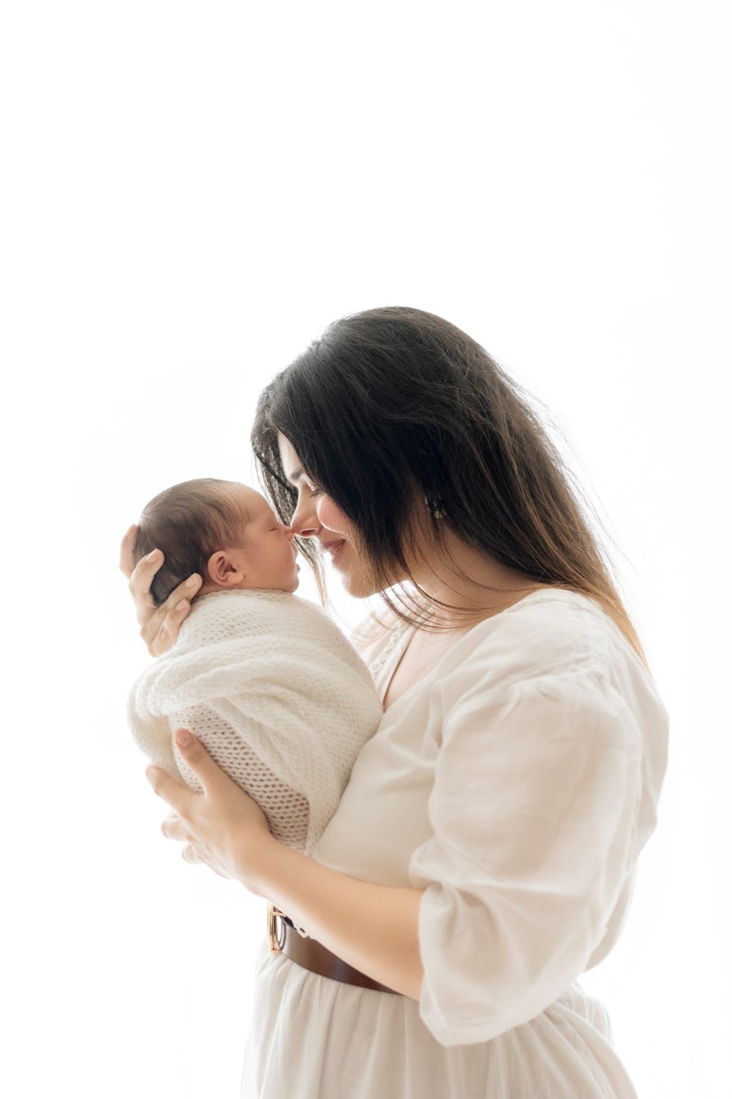 Tender moment between mother and newborn baby wrapped in white against light background.