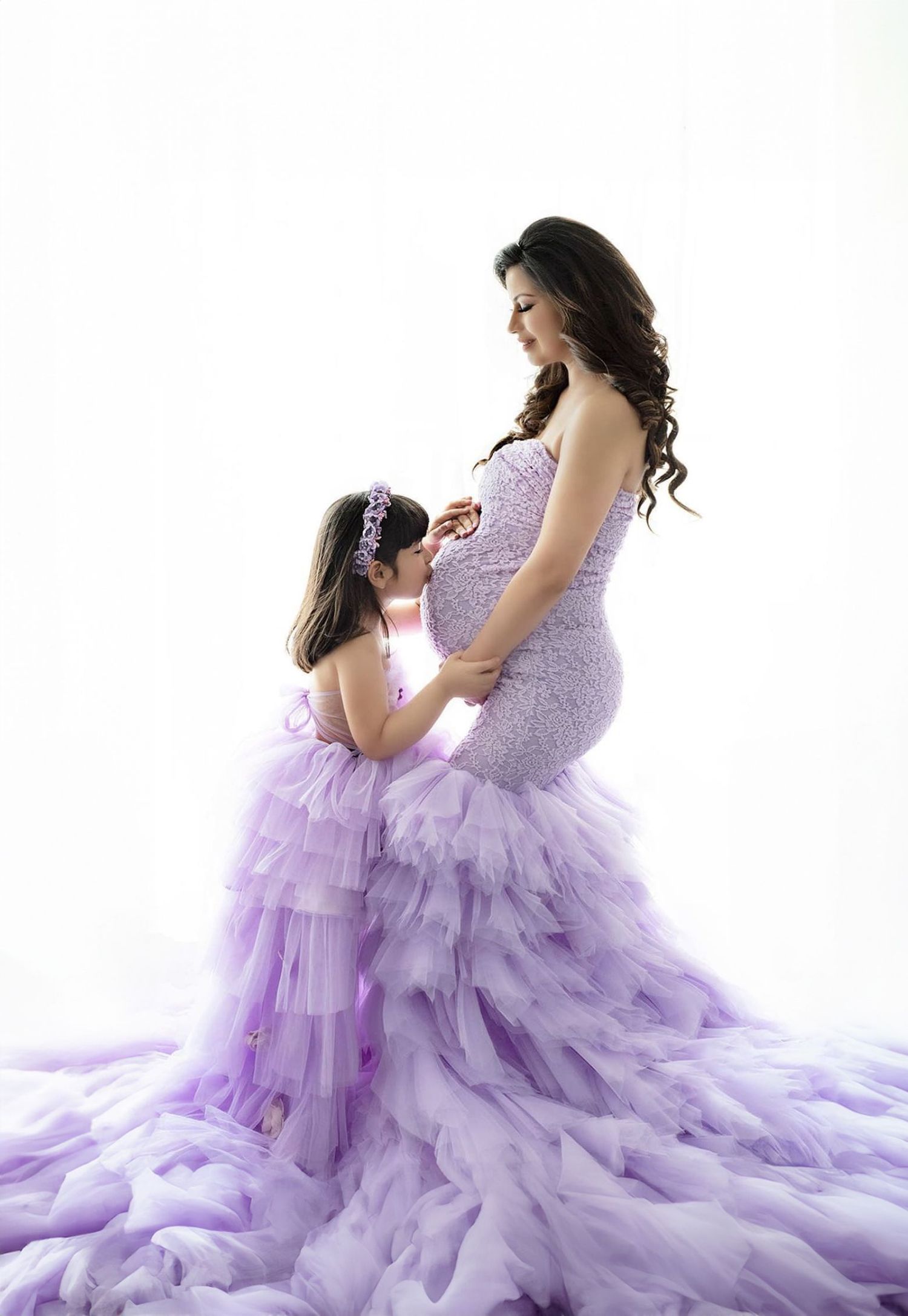 Sweet maternity moment in matching lavender tulle gowns with ruffled details against white backdrop.