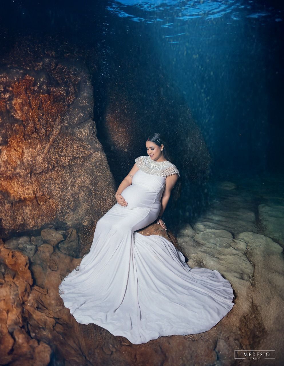 Dramatic underwater maternity photo featuring white gown against rocky cave backdrop with blue water.