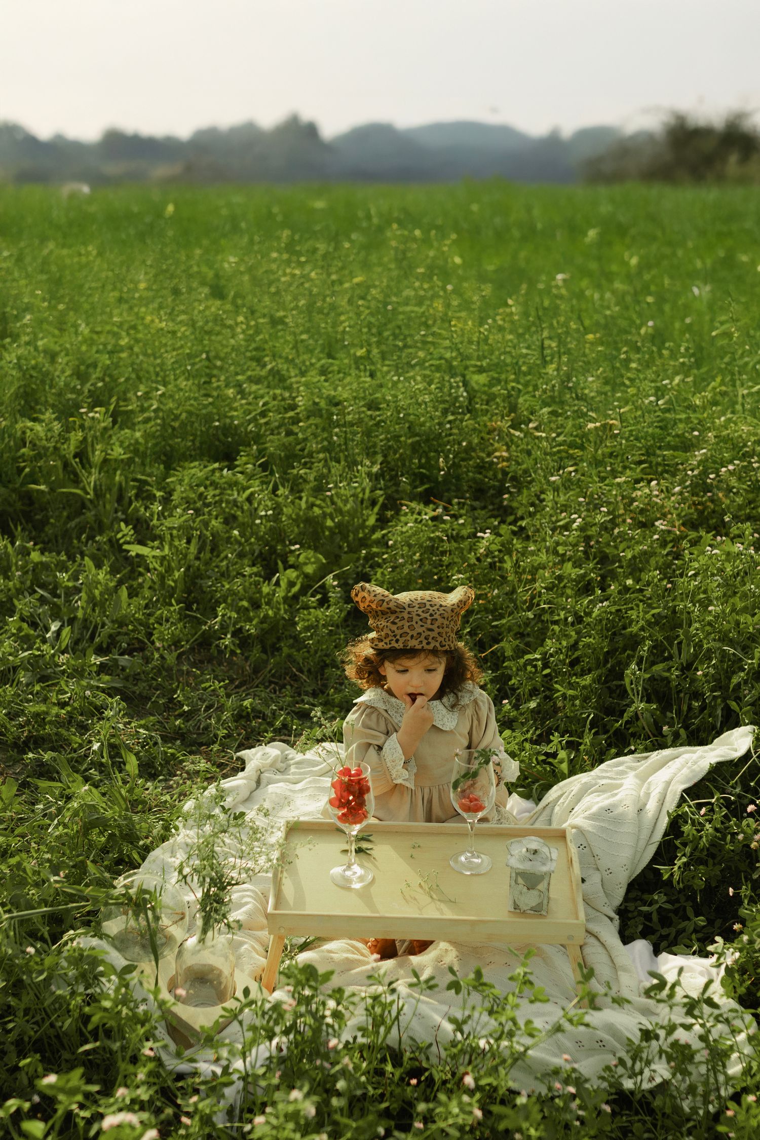 Young child in bear hat enjoys a picnic on white blanket in lush green field with tray of drinks.