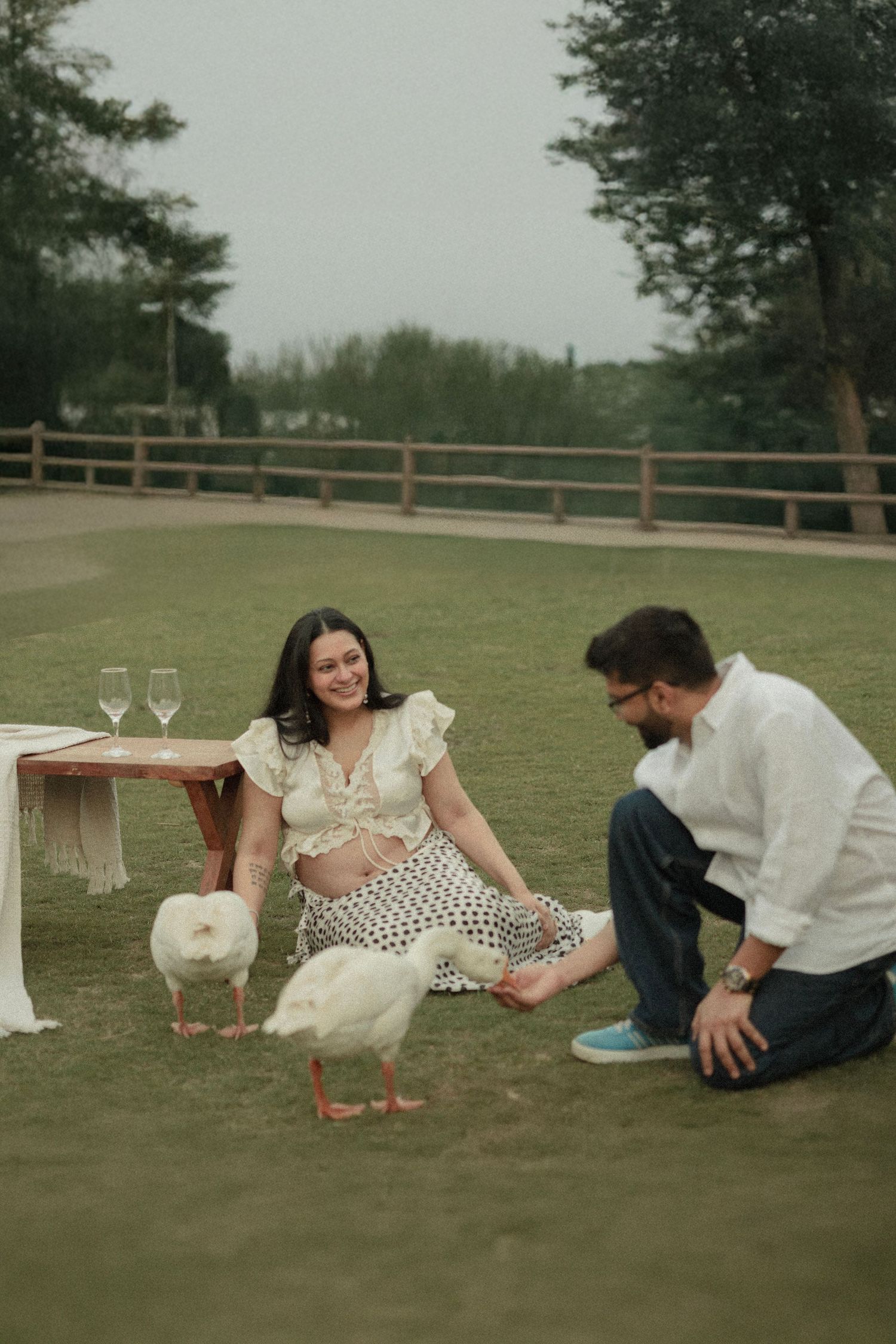 Pregnant woman and man sitting on grass with white ducks during an outdoor maternity photoshoot.