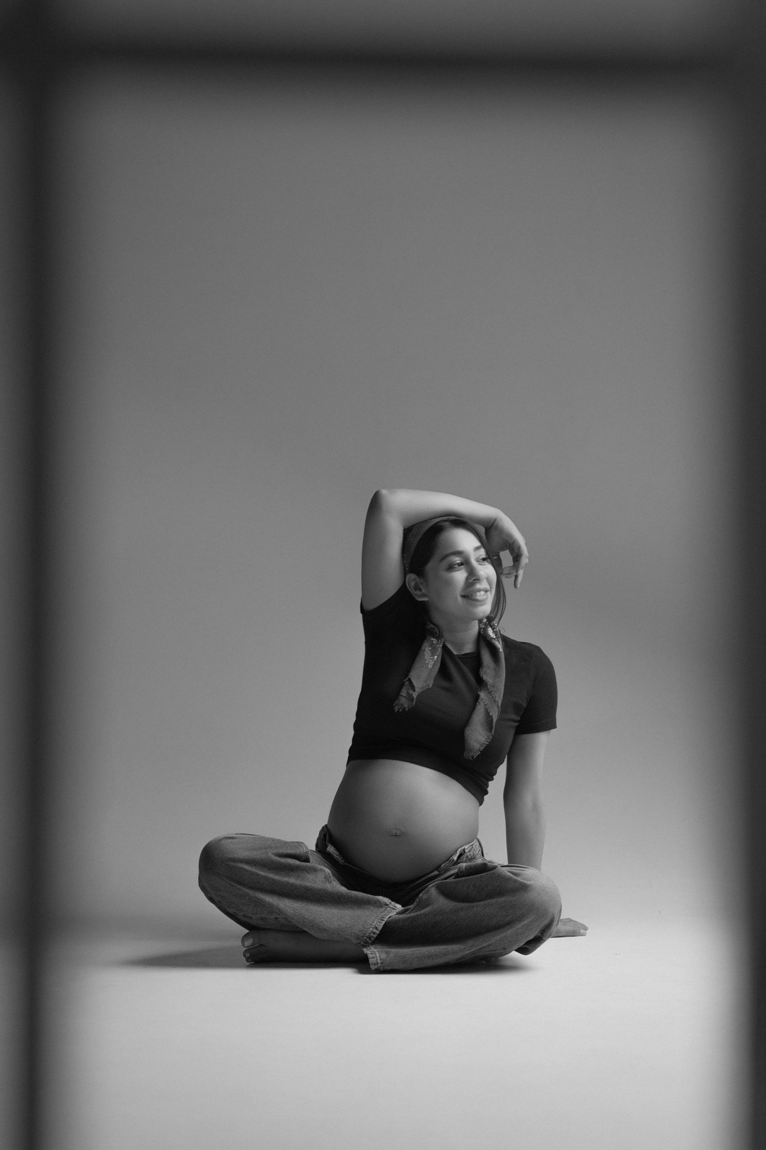Black and white photo of a pregnant woman sitting cross-legged on the floor with her arm raised above her head.