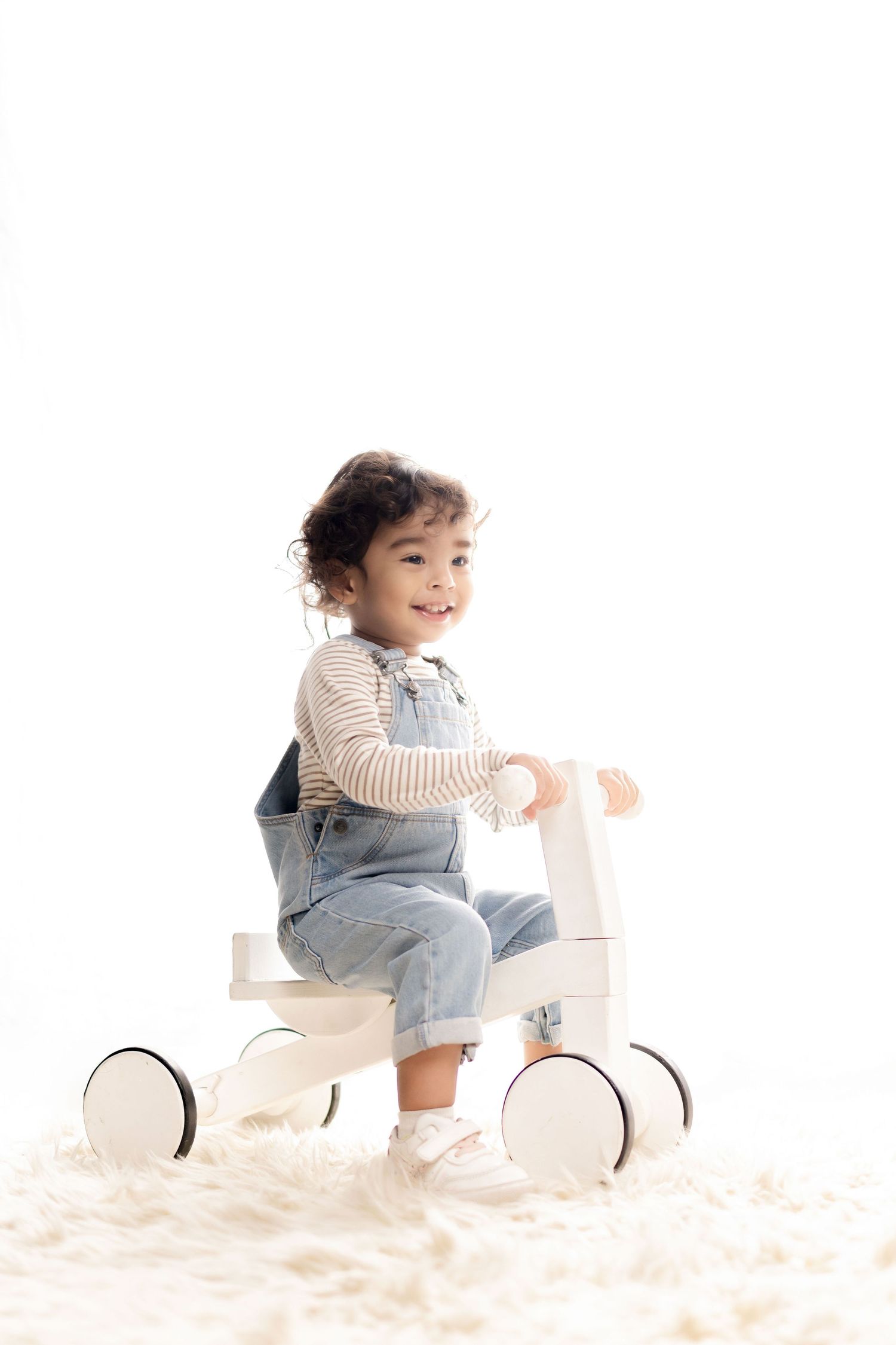 Smiling toddler girl in denim overalls riding a small wooden balance bike on a fluffy white rug.