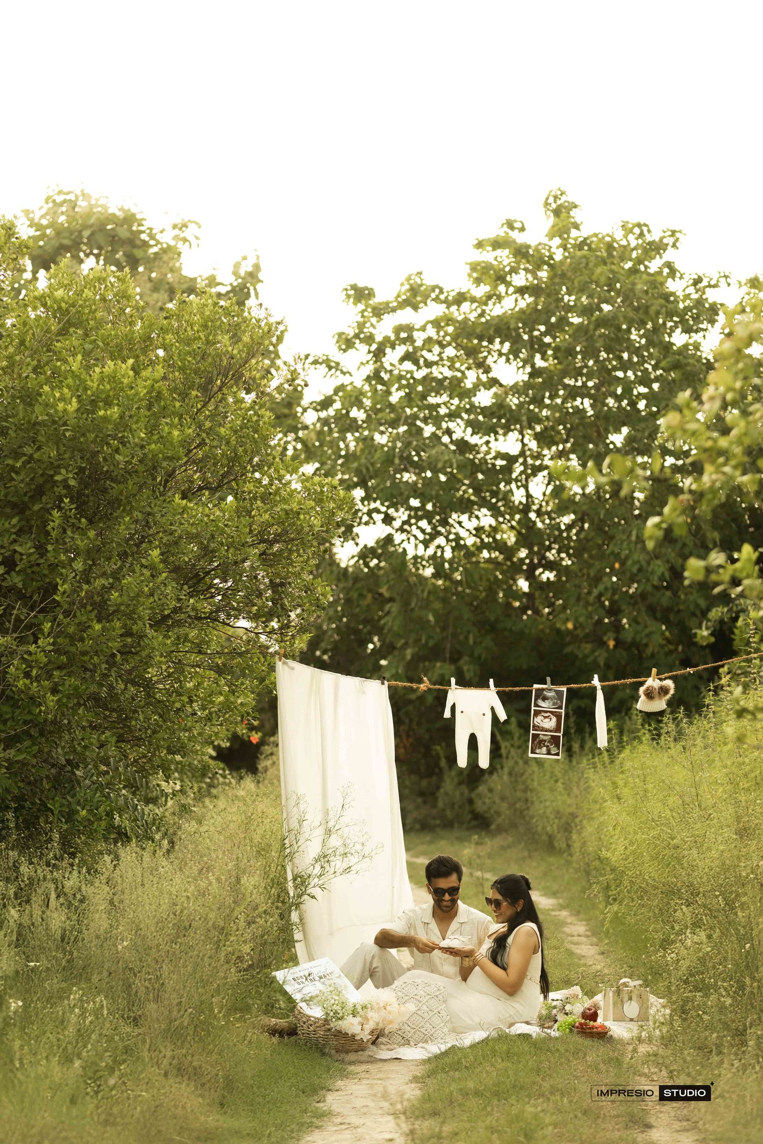 Couple sitting on grass in nature with white sheet backdrop and baby clothes hanging on a line.