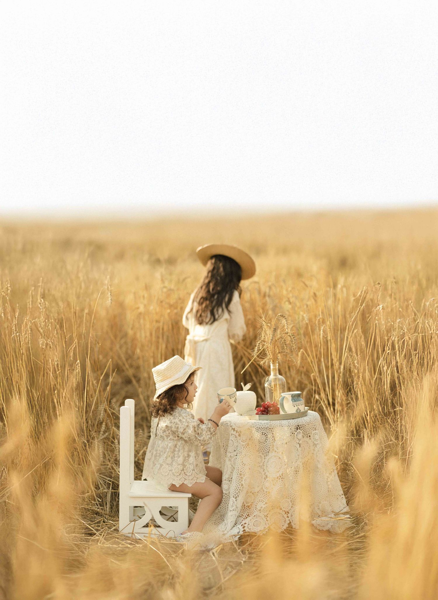 Two girls in white dresses enjoy a tea party setup in a golden wheat field.