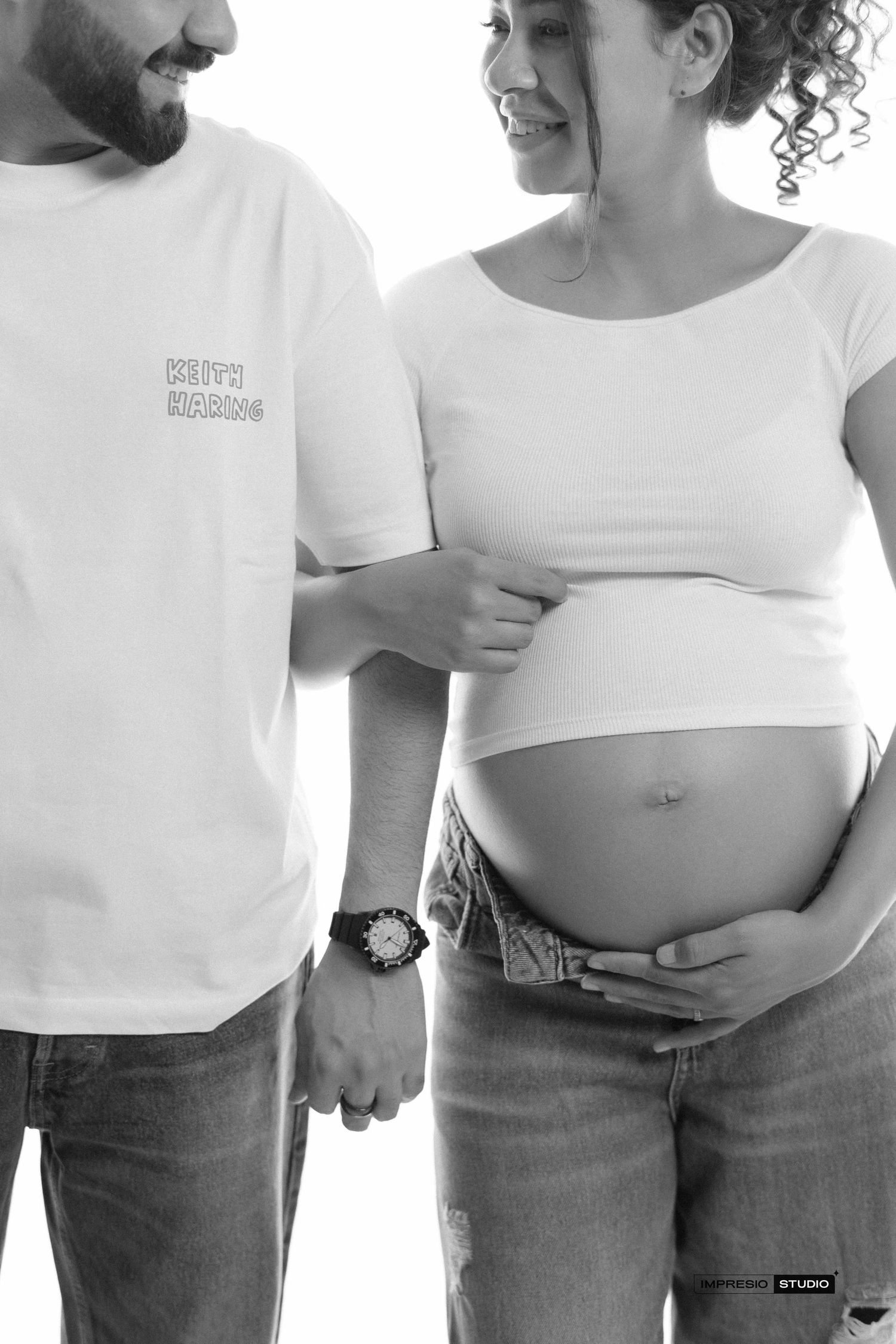 Black and white maternity photo of a couple in white shirts and jeans, woman cradling pregnant belly in studio.