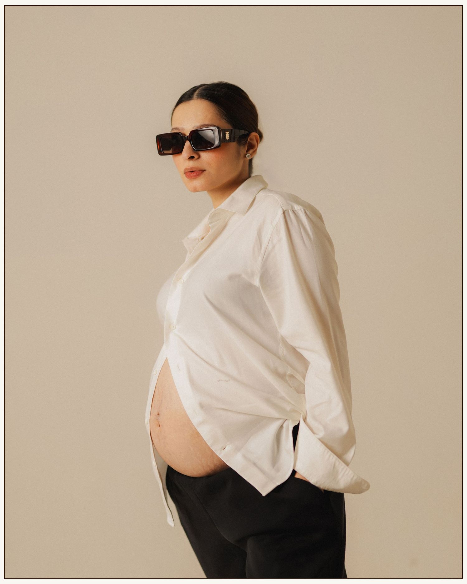 Pregnant woman in white oversized shirt and black sunglasses posing against a neutral beige background.