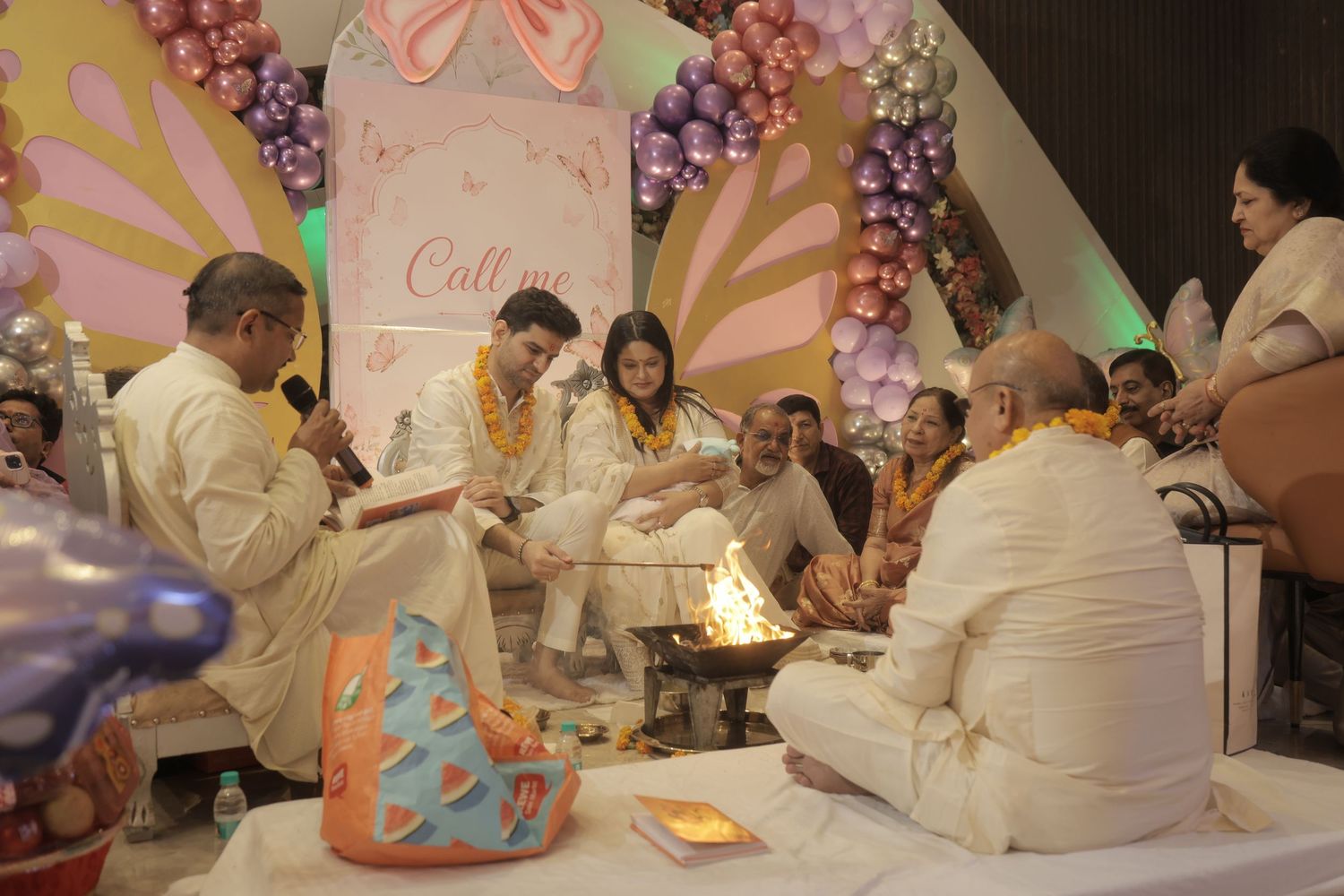Family and guests in white traditional attire sit around a sacred fire during a Hindu wedding ceremony decorated with colorful balloons.