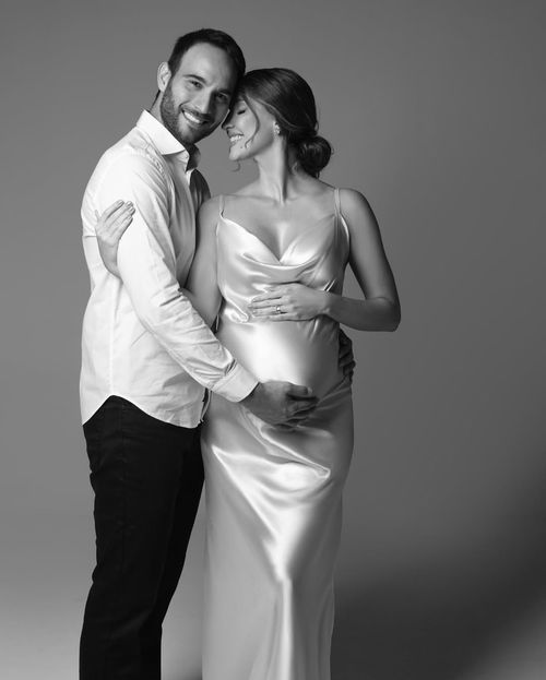 A couple shares an intimate black and white moment, dressed elegantly in white attire against a simple studio backdrop.