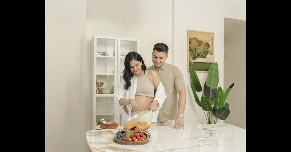 A couple prepares food together in a bright modern kitchen with white cabinets and a decorative plant for maternity shoot.