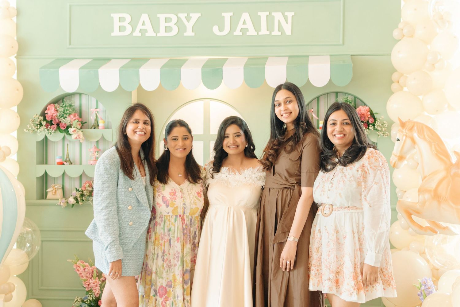 Five women smiling at a Baby Jain baby shower in front of a green decorative backdrop with flowers.