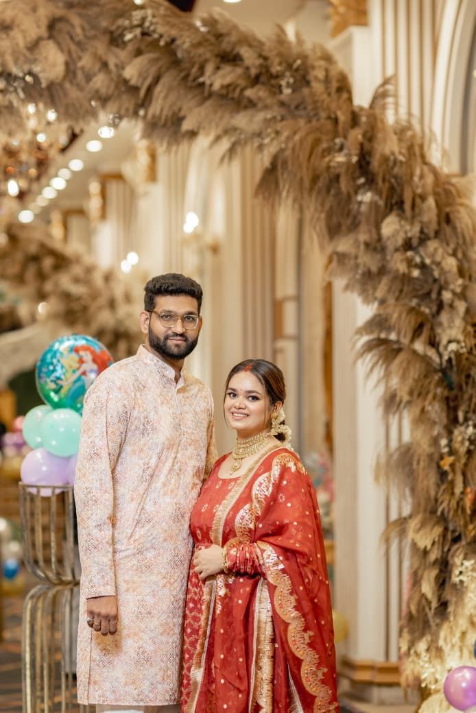 A couple in traditional South Asian attire poses elegantly at a decorated venue with pampas grass and colorful balloons.